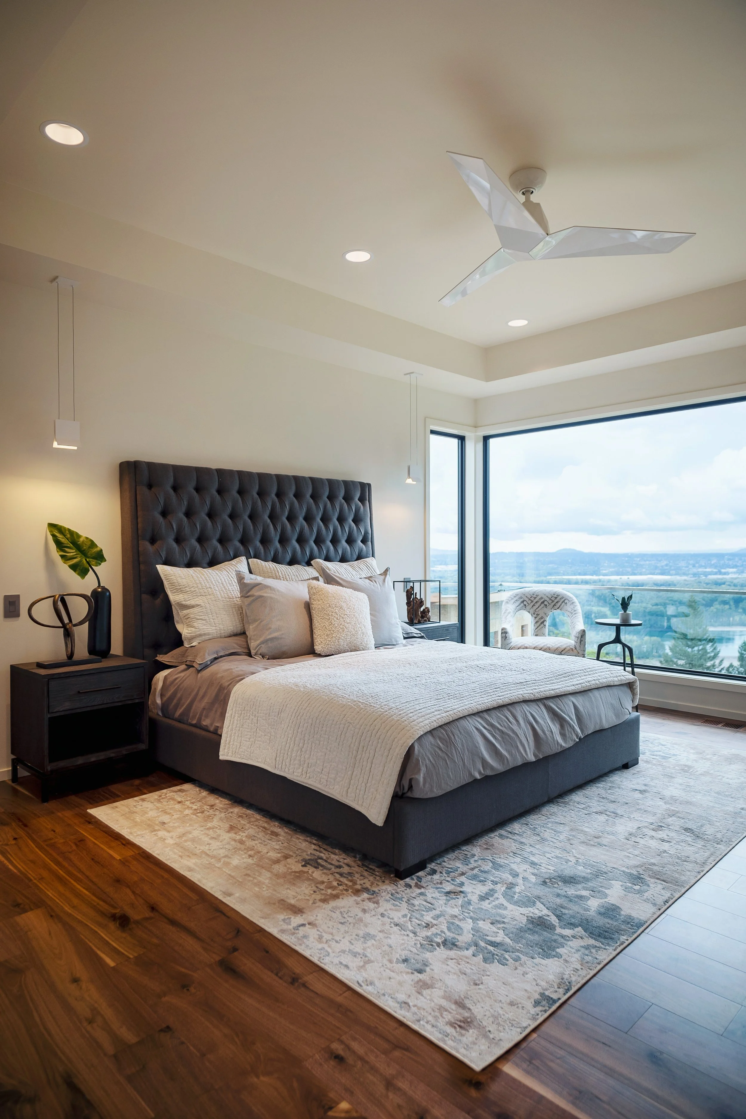 Modern bedroom with large bed, tufted headboard, nightstand, side tables, and a window view of a cityscape and sky.
