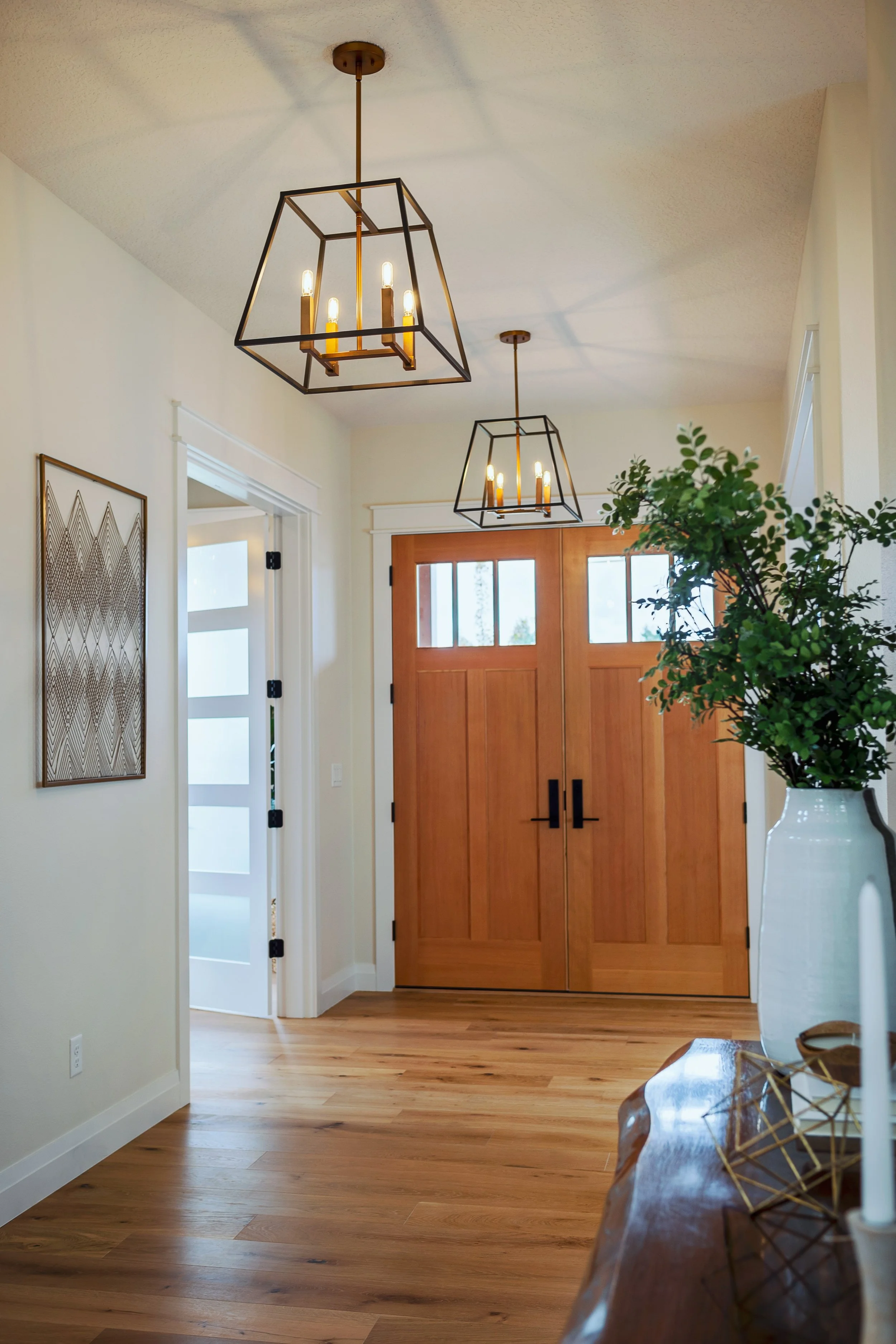 Foyer with wooden front door, pendant light fixtures on the ceiling, a white wall with a decorative art piece, hardwood flooring, and a large potted green plant in a white vase.