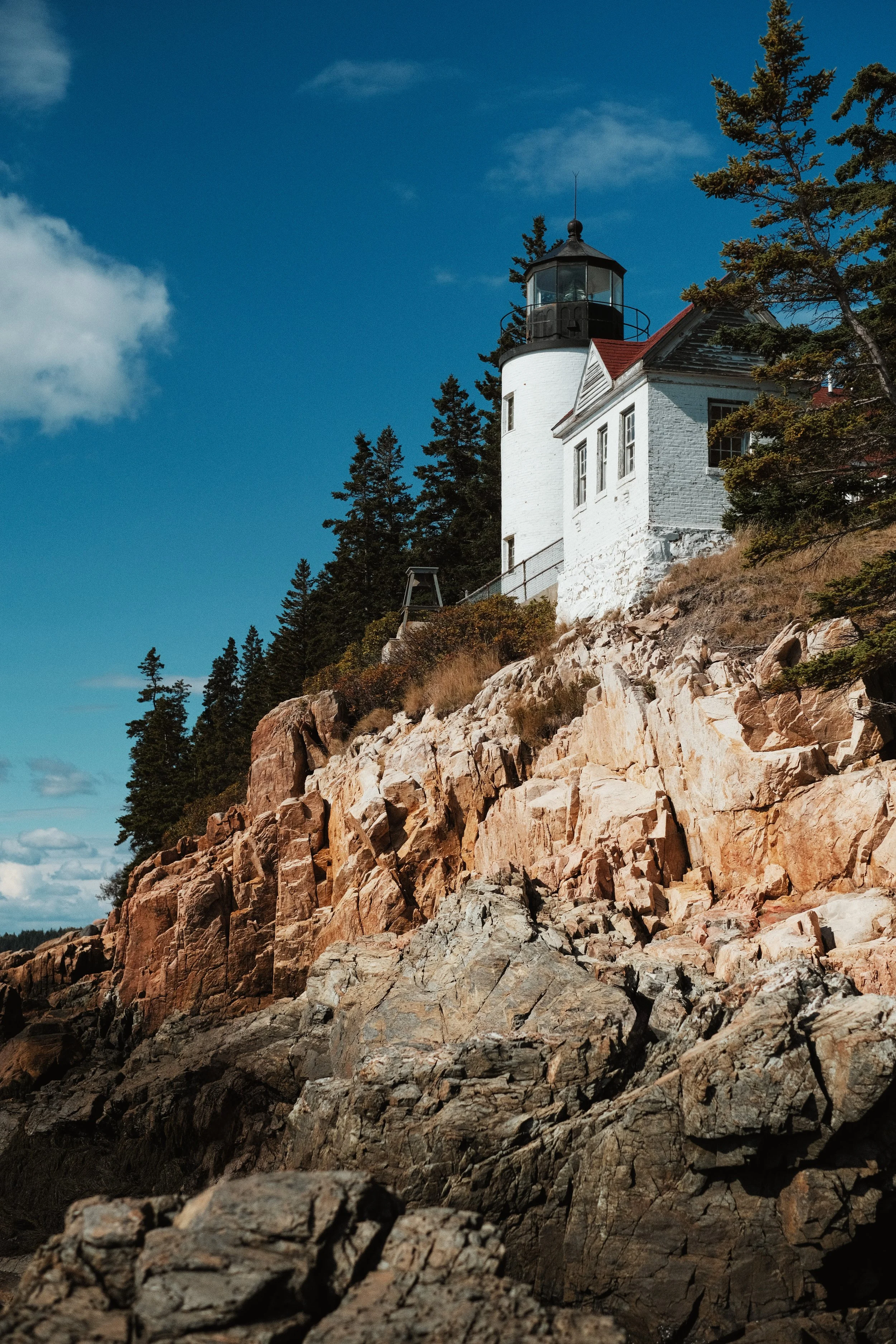 A lighthouse on a rocky hillside surrounded by trees, with a clear blue sky in the background.