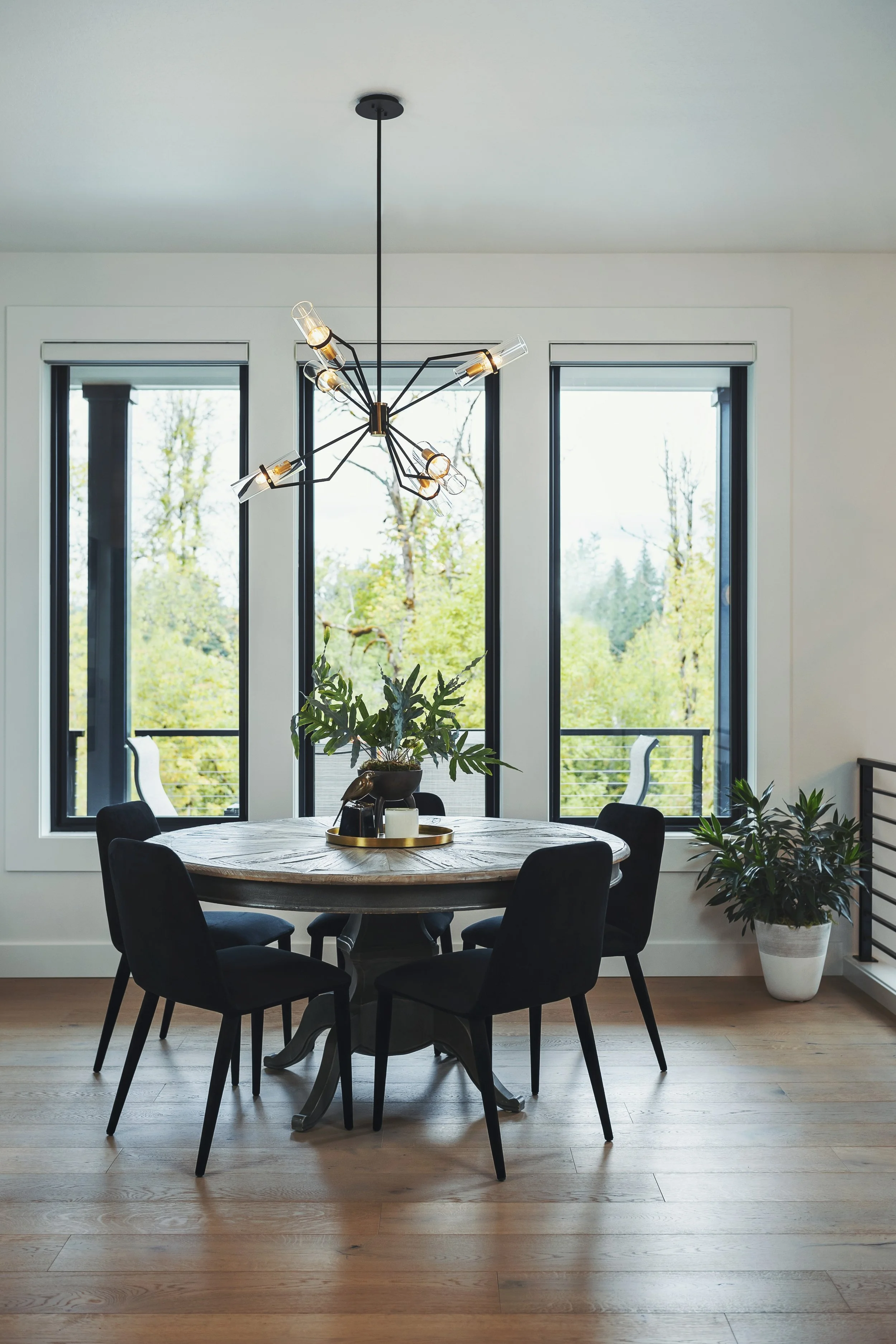 A dining room with a round table, six black chairs, a modern chandelier, potted plant in a white pot, and large windows showing green trees outside.