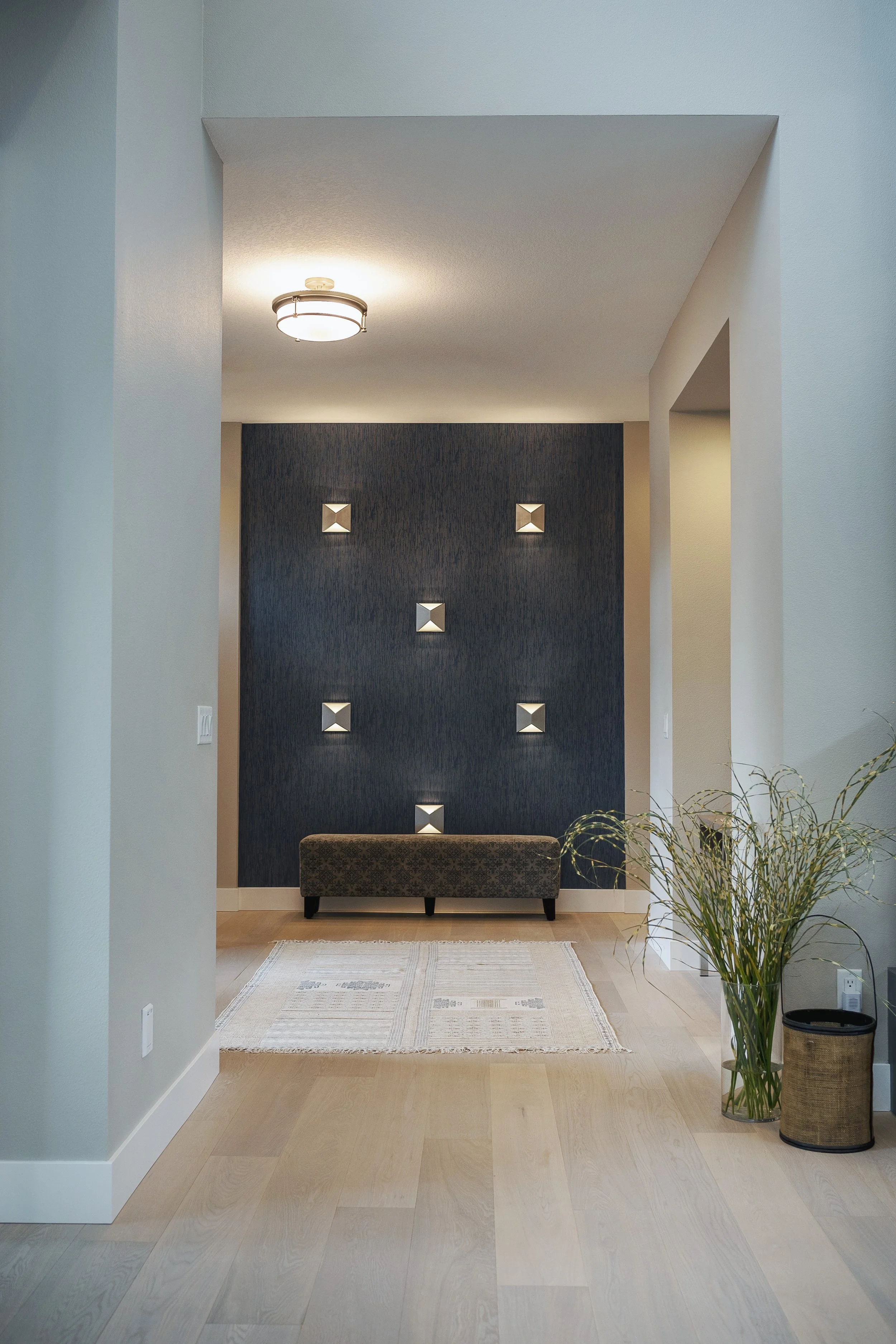Interior entryway with a dark textured accent wall featuring decorative wall art, a small upholstered bench, a patterned rug, a tall glass vase with a leafy plant, and a woven basket, with a light-colored hardwood floor and a minimalist ceiling light