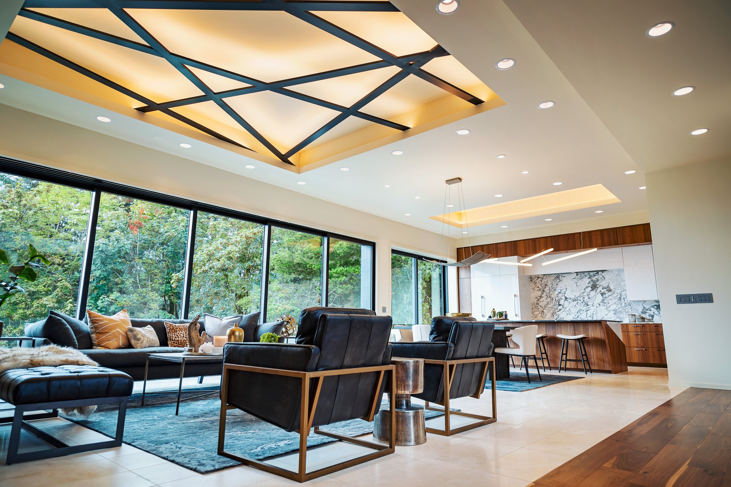 Modern living room with black leather sofa, patterned throw pillows, large windows showing green trees outside, a marble kitchen backsplash, and a wooden bar or counter with stools.