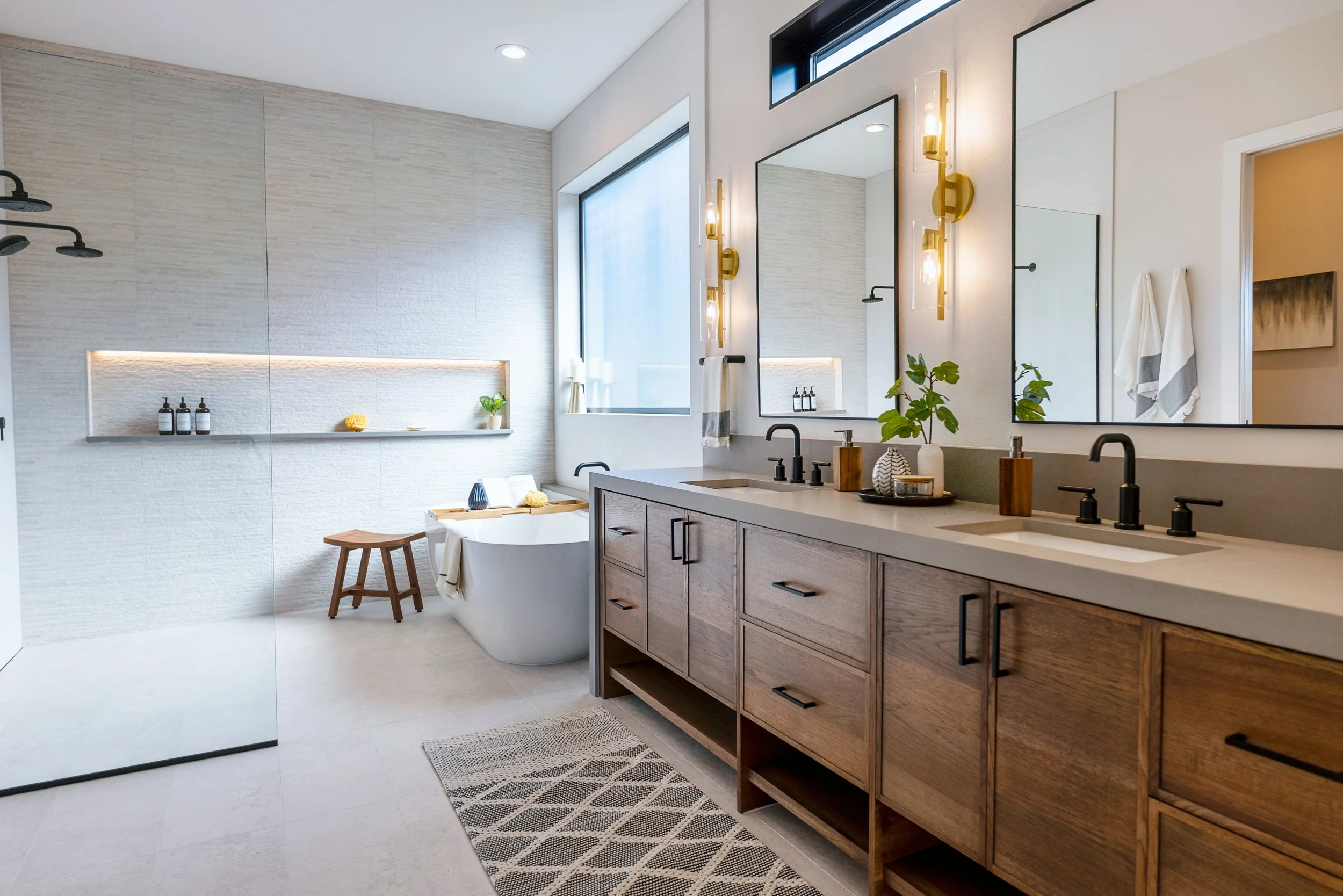 Modern bathroom with a walk-in shower, bathtub, large vanity with dual sinks, and contemporary decor. Features include black fixtures, wooden cabinetry, large mirrors, wall-mounted lighting, and natural light from high windows.