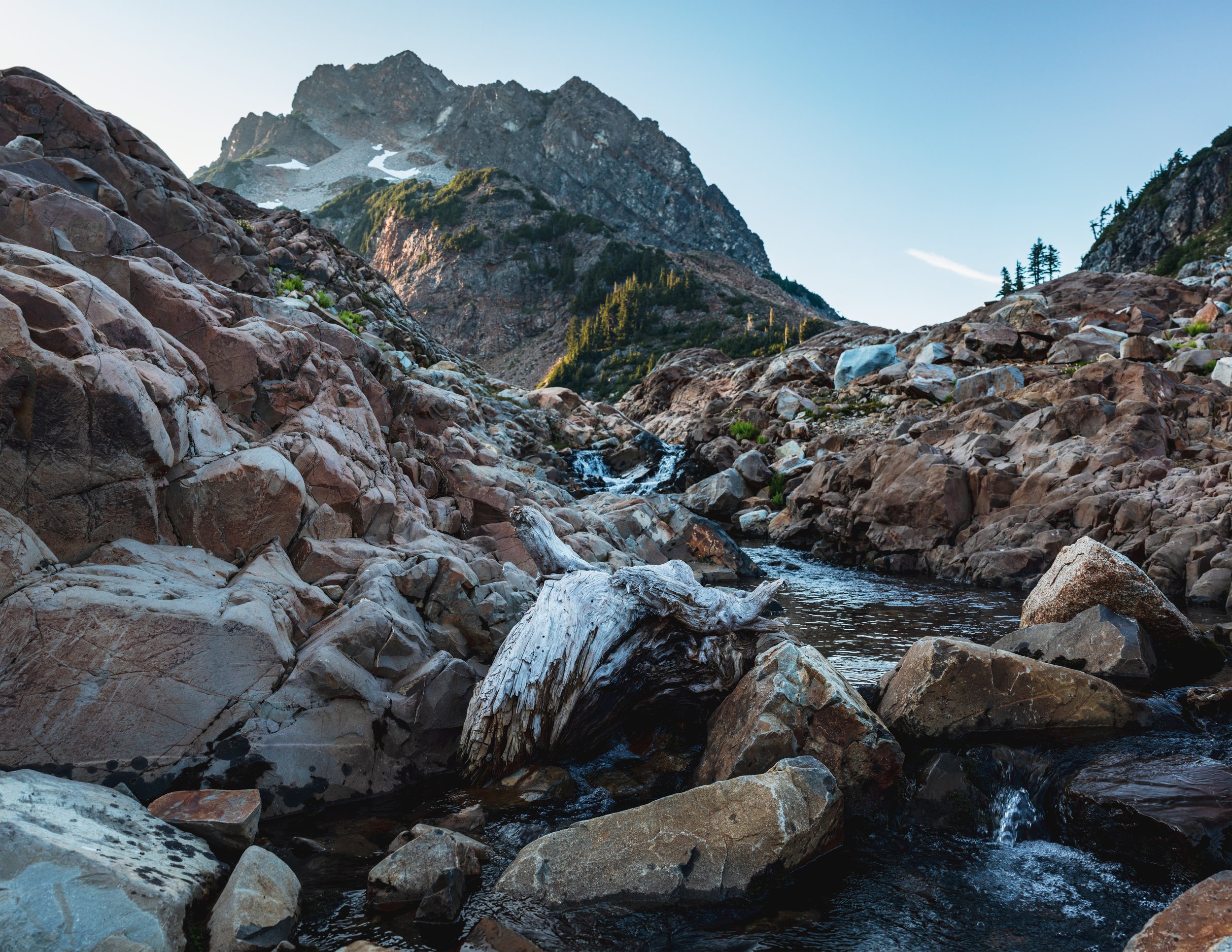 A rocky mountain stream flowing through a valley with large boulders and green trees, with snow-capped peaks in the background against a clear blue sky.