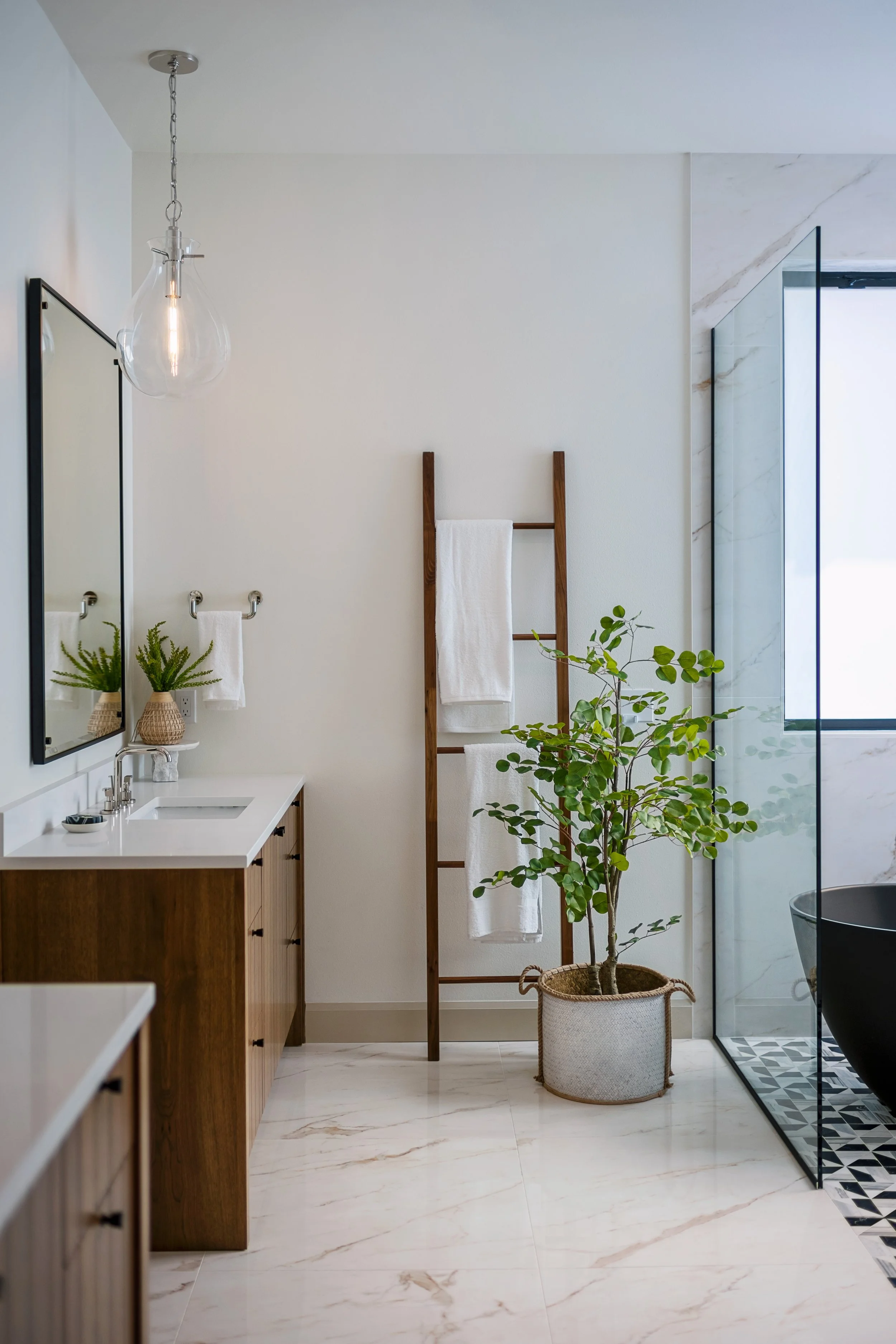 Modern bathroom with wooden vanity, white countertop, large mirror, hanging glass pendant light, potted plants, ladder towel rack, and glass shower with patterned black-and-white tiles.