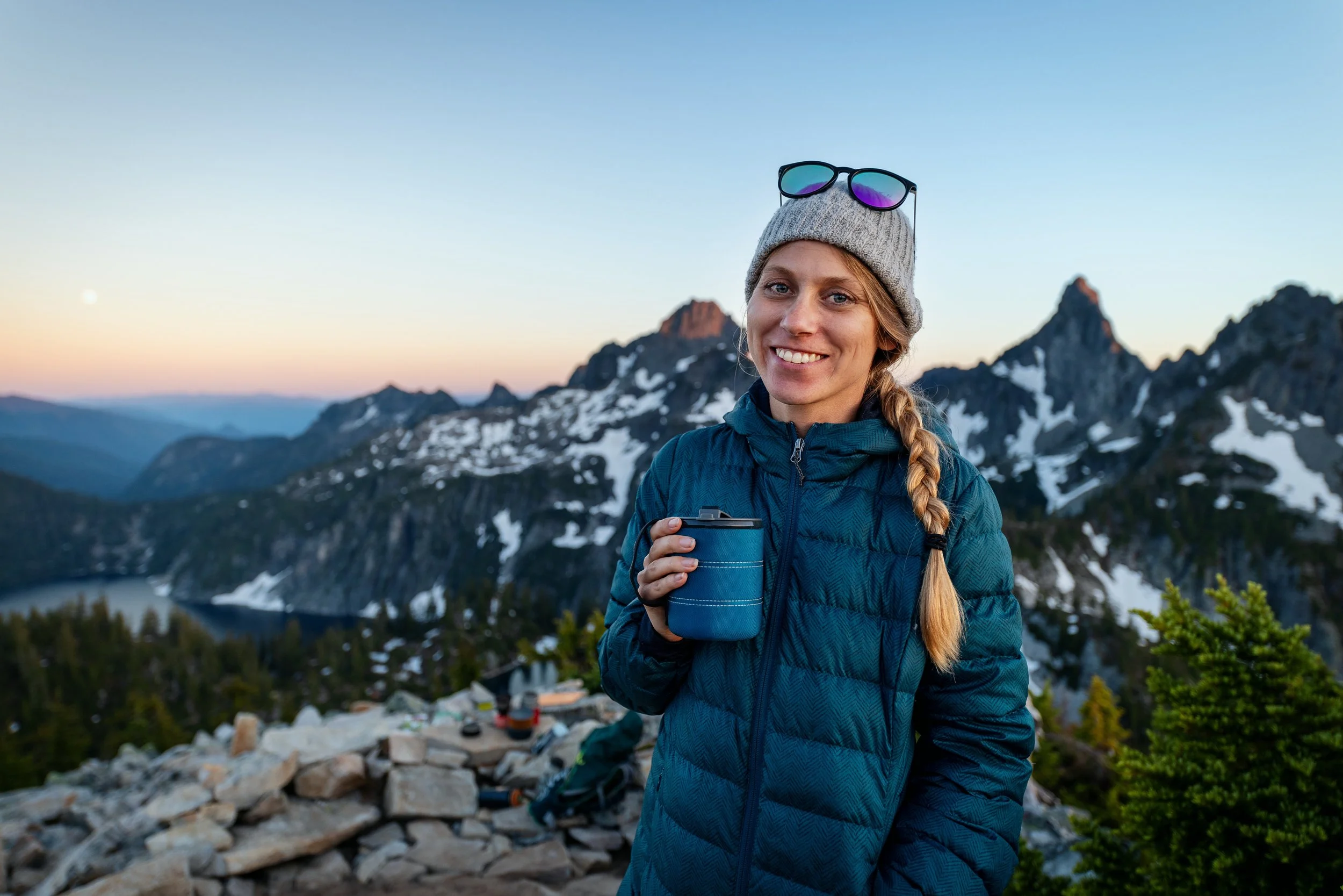 A woman with long blond hair in a braid holding a travel mug, standing outdoors near a mountain lake during sunset, with snow-capped peaks in the background and camping gear nearby.