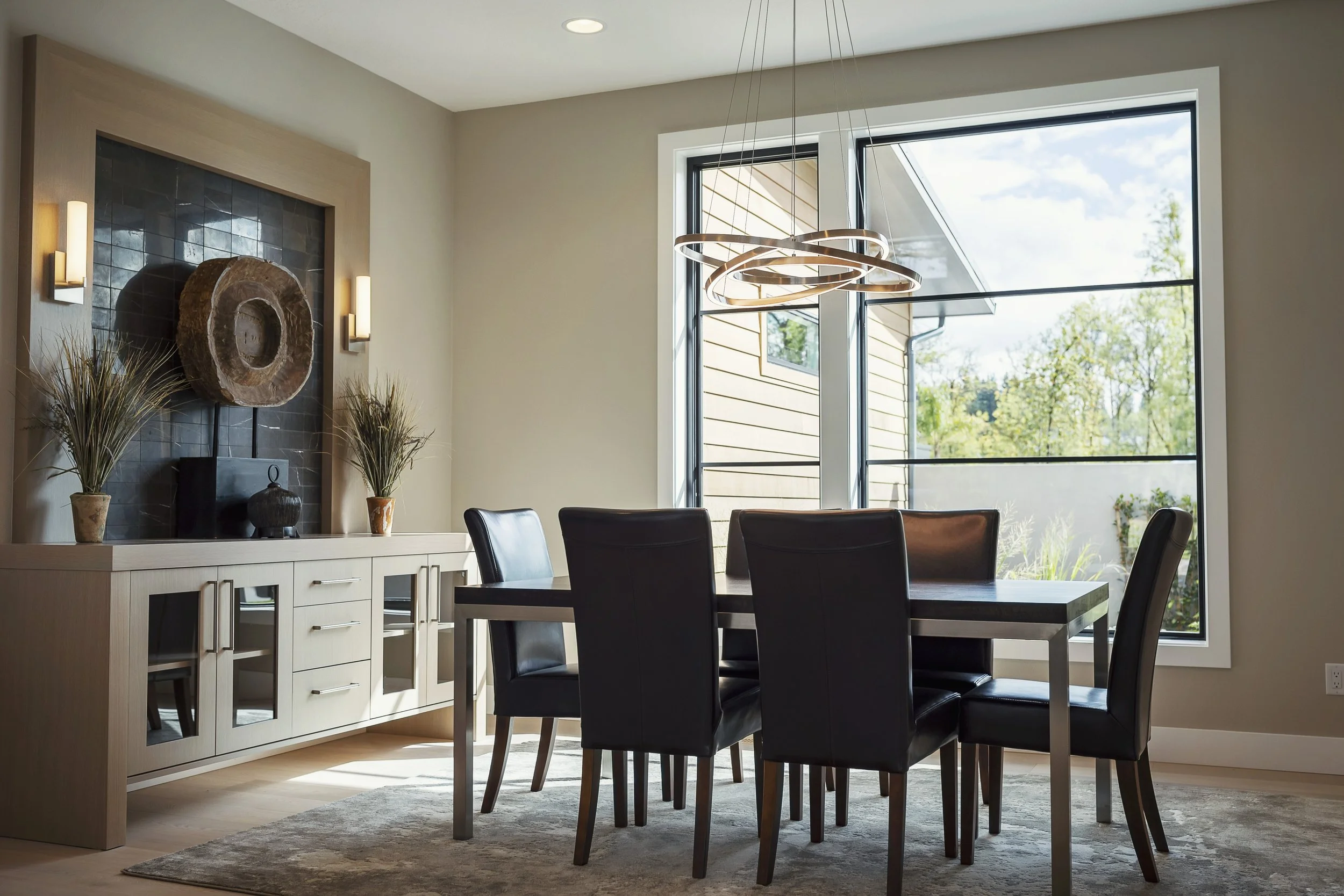 A modern dining room with a light-colored wooden sideboard, black chairs around a matching wooden table, a large window with a view of the outdoors, a contemporary circular chandelier, and decorative vases with plants on the sideboard.