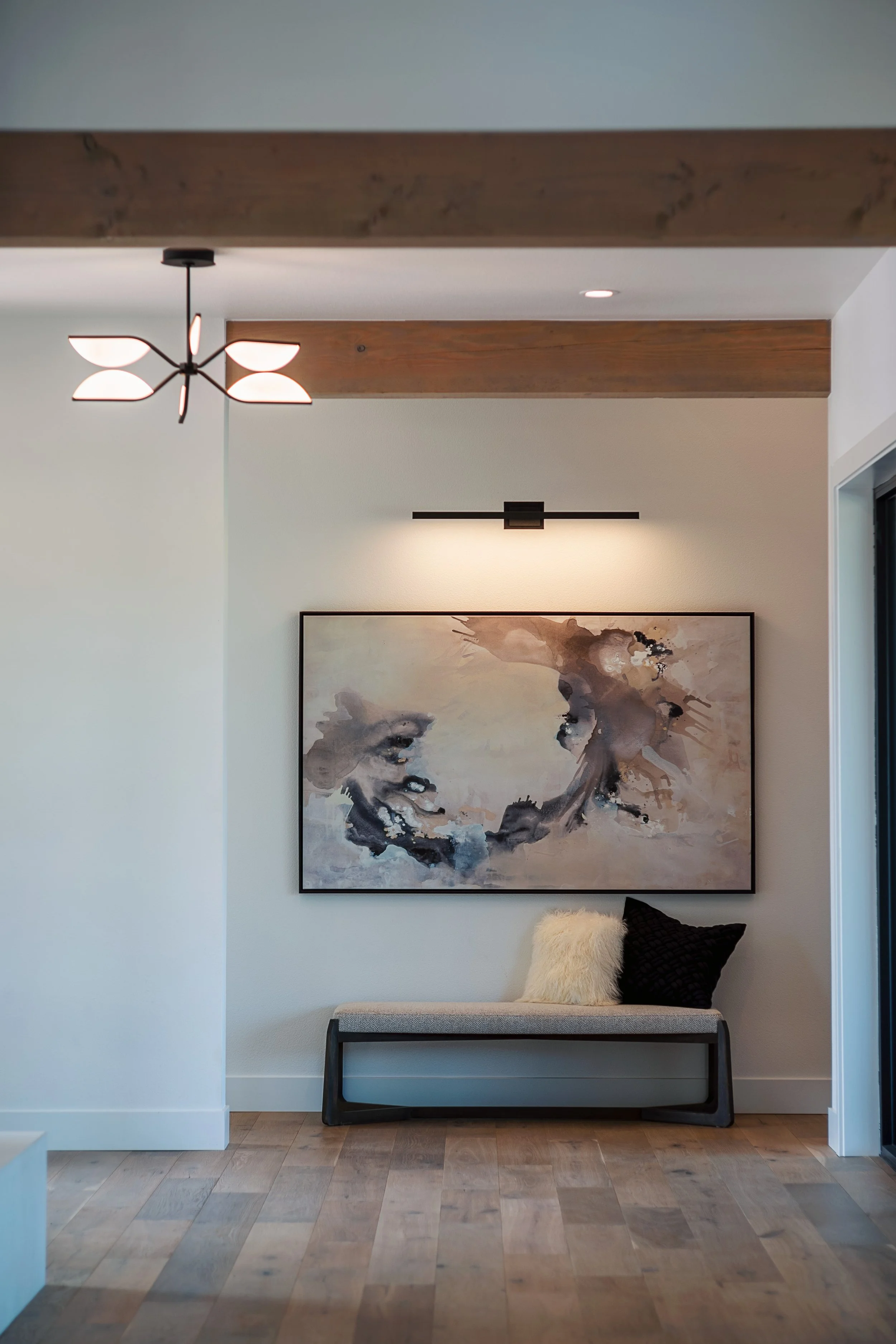 A modern interior with a wall art, a small bench with pillows, wooden ceiling beams, and ceiling lights.
