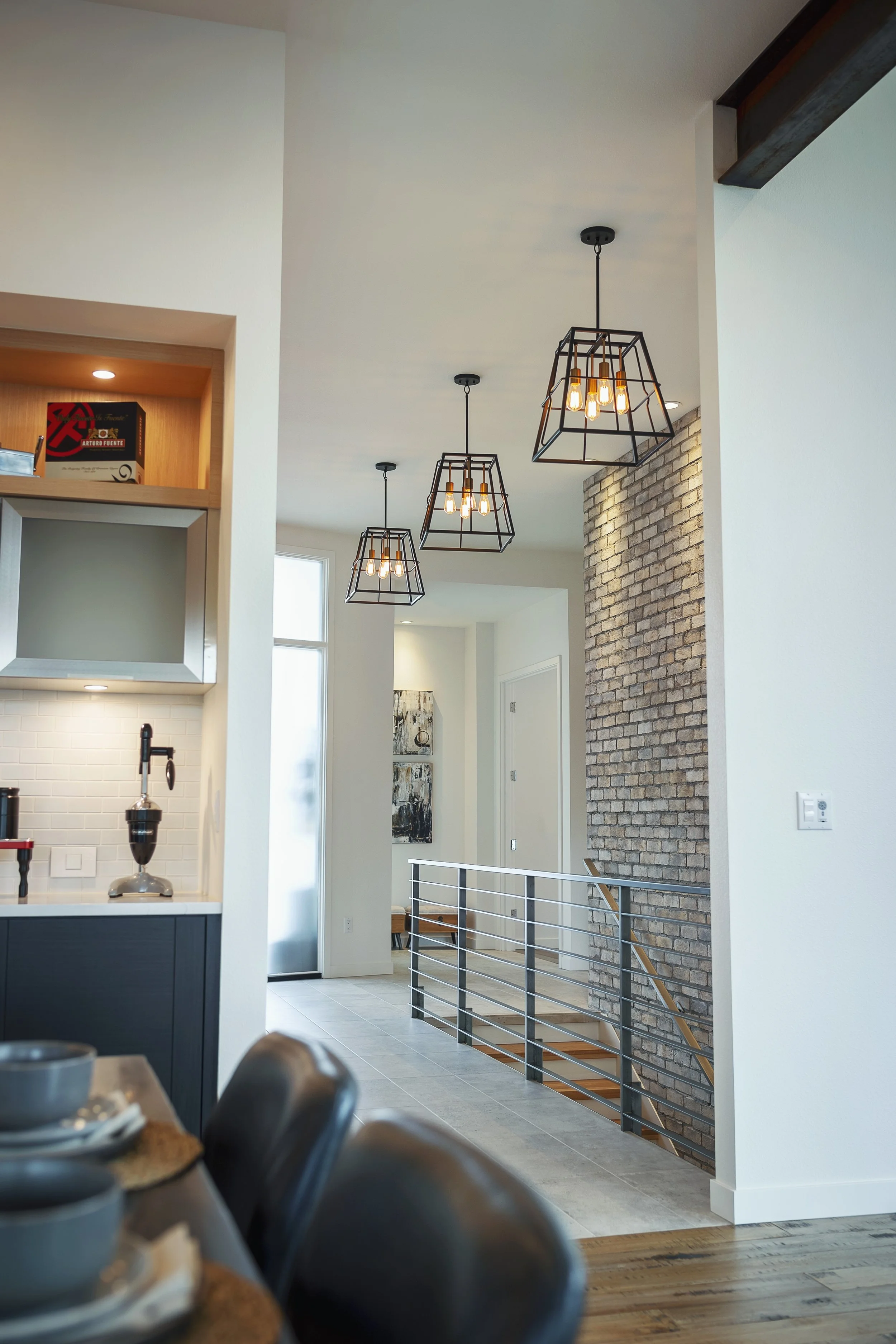 Interior view of a modern home with hanging pendant lights, a brick accent wall, and a staircase with metal and wood railing.
