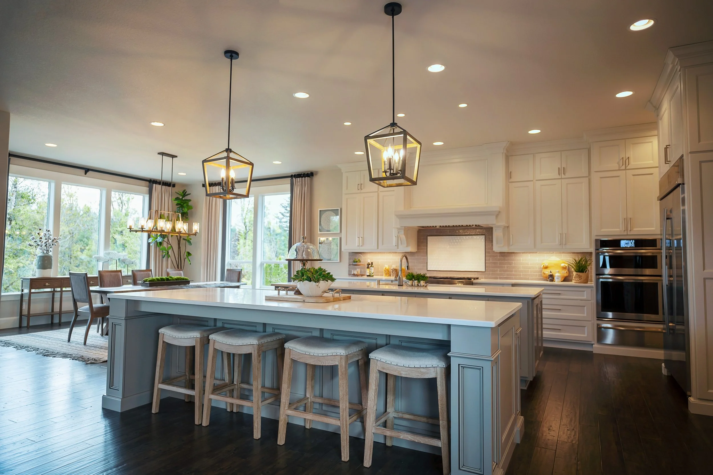 Bright kitchen with white cabinetry, a large island with barstools, pendant lights, and large windows, overlooking a dining area with a chandelier and outdoors greenery.