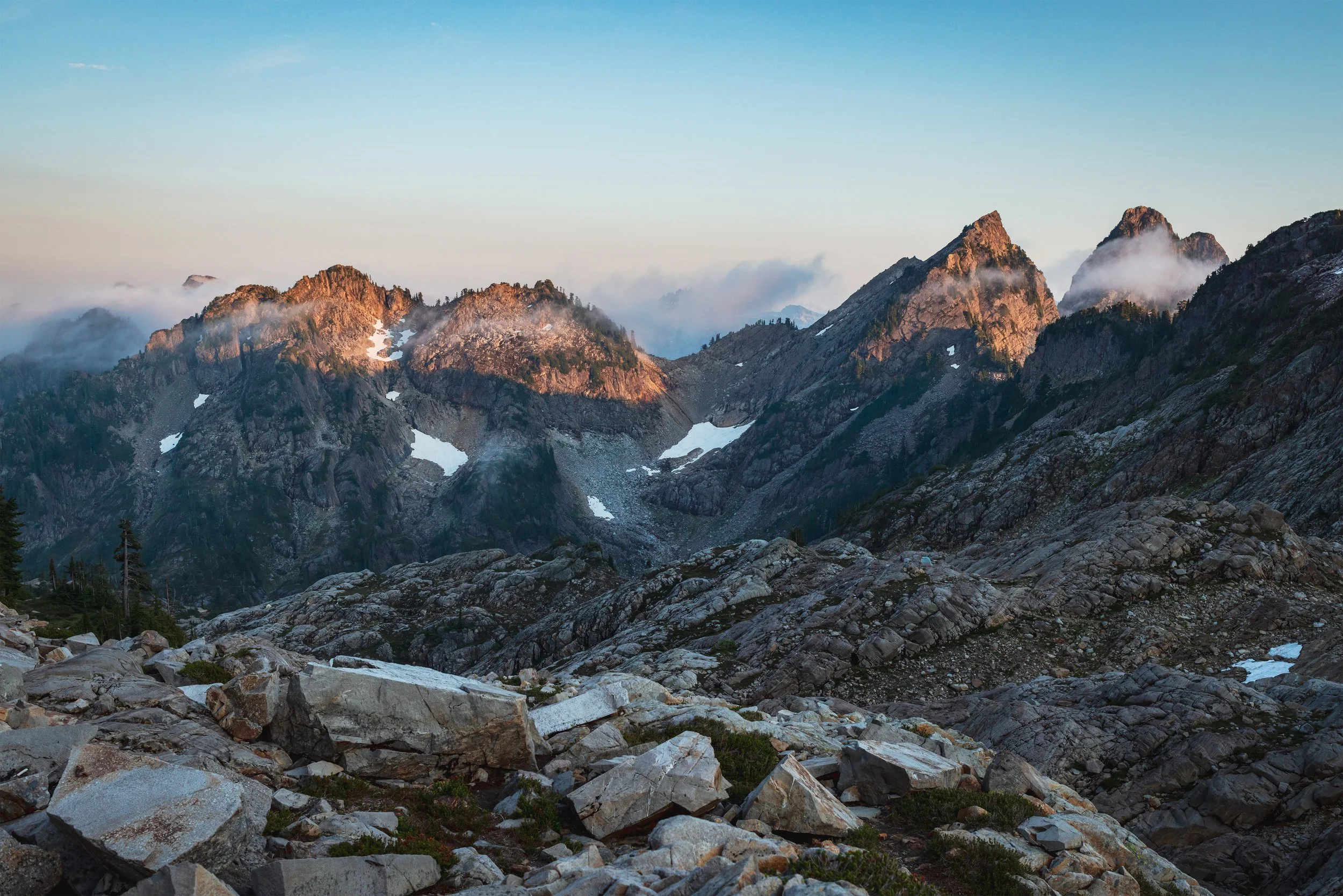 Mountain range with rugged peaks partially covered in snow, shrouded in clouds, under a clear blue sky.