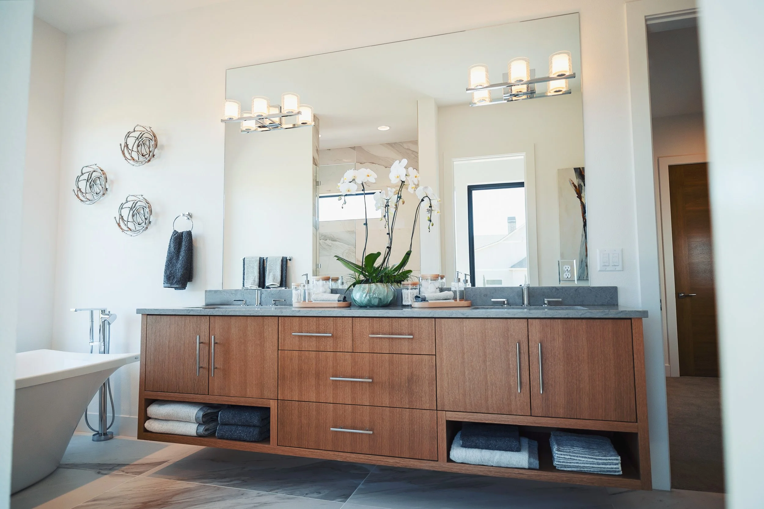 Modern bathroom with wooden vanity, white flowers in a pot, large mirror, and decorative wall art, with natural light coming through the window.