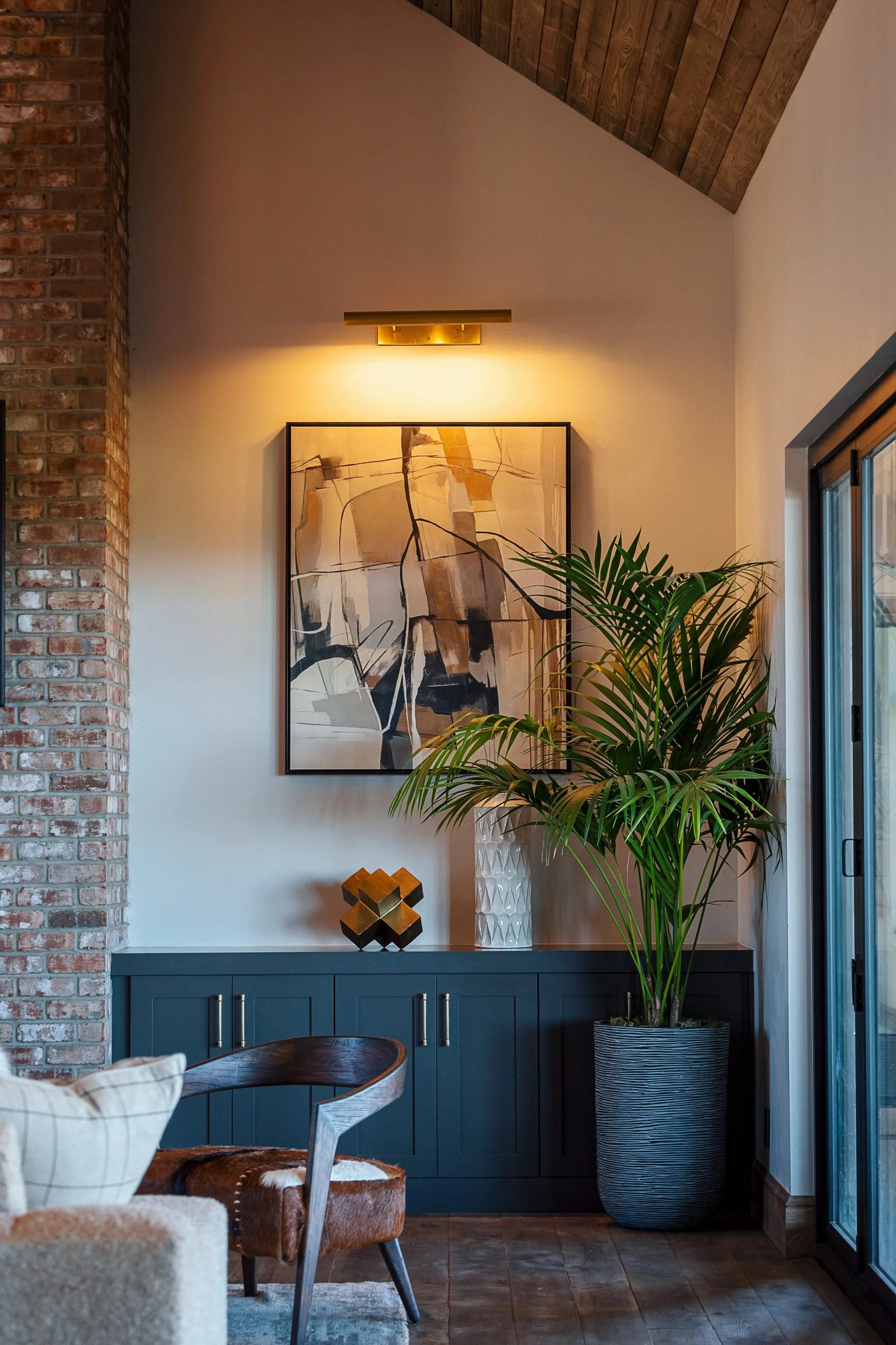 Living room corner with brick and wood wall, abstract art on the wall, large potted plant, blue cabinet, and decorative objects.