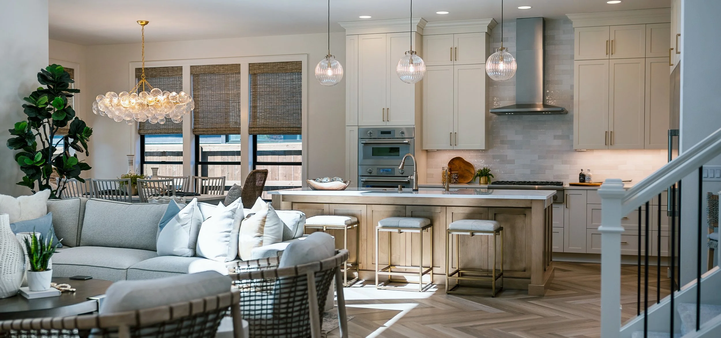 Open-concept living room and kitchen with white cabinetry, a kitchen island with bar stools, modern pendant lights, a large window with woven shades, and a beige sectional sofa with throw pillows.