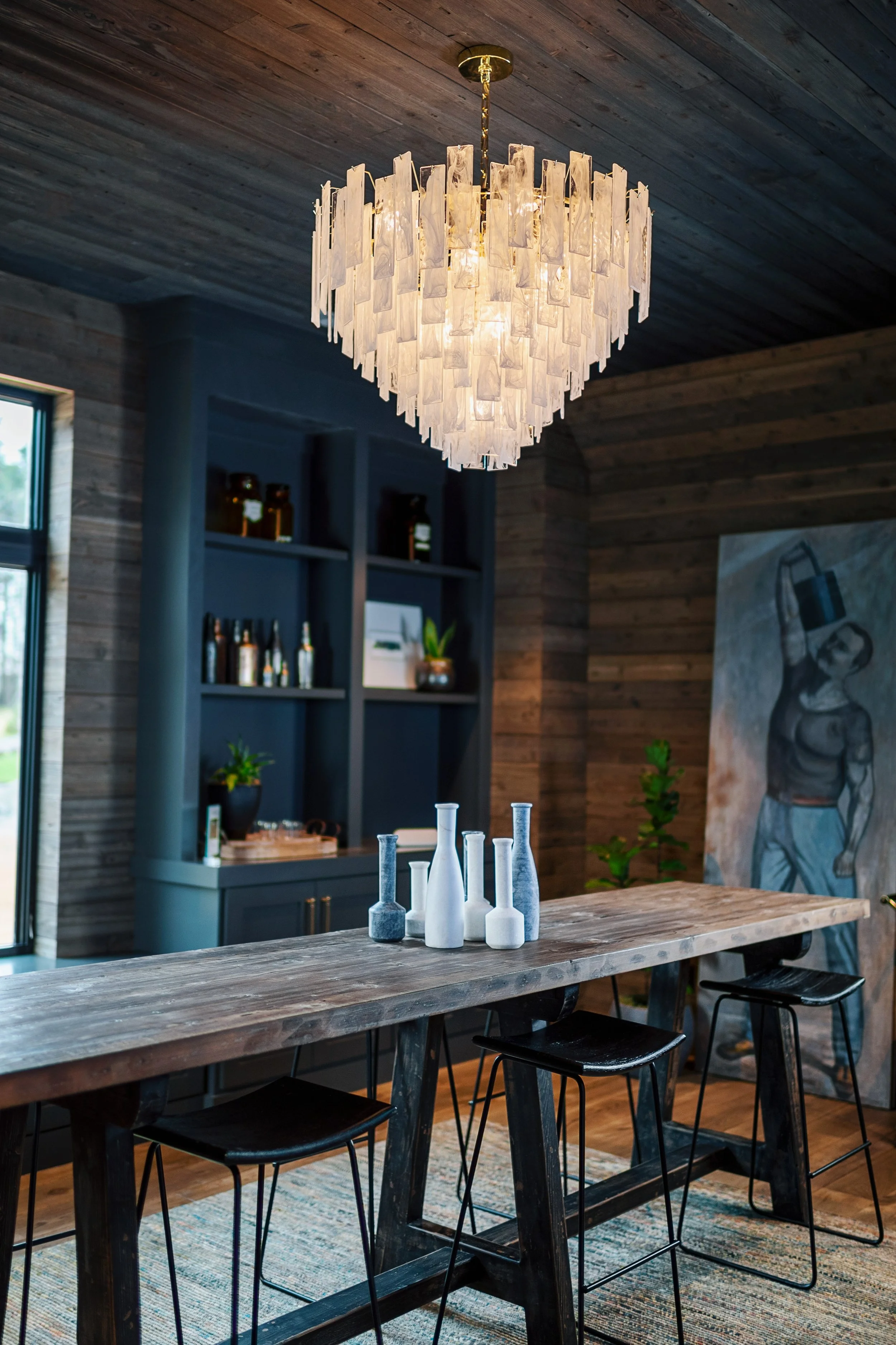 A rustic dining room with a large wooden table. White and gray vases are arranged on the table. The room has wooden walls and a wooden ceiling. A chandelier with white decorative panels hangs above the table. There is a built-in dark blue cabinet wit
