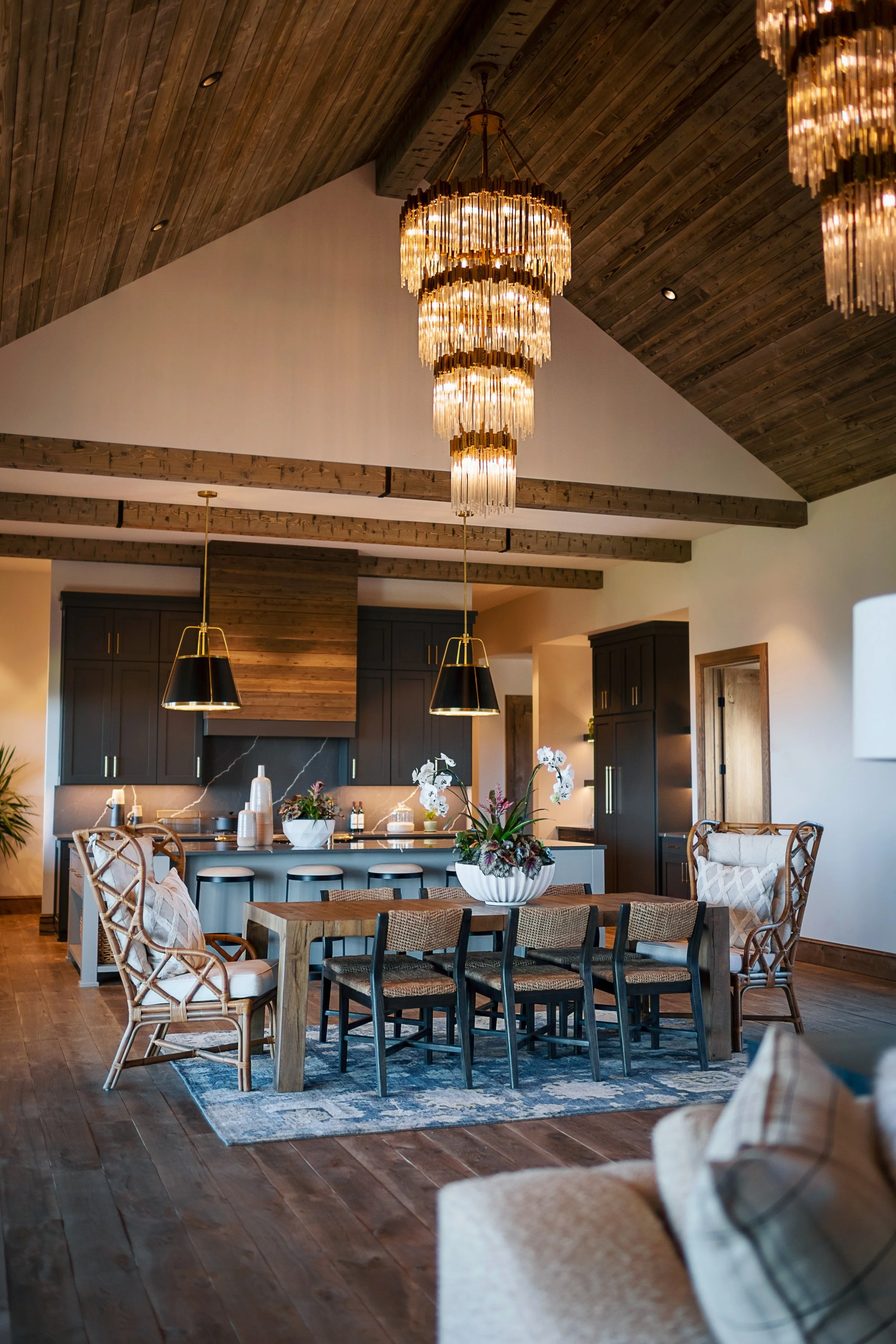 Living room with a wooden ceiling and chandelier, open kitchen with dark cabinets, wooden dining table with chairs, floral centerpiece, and a cozy seating area.
