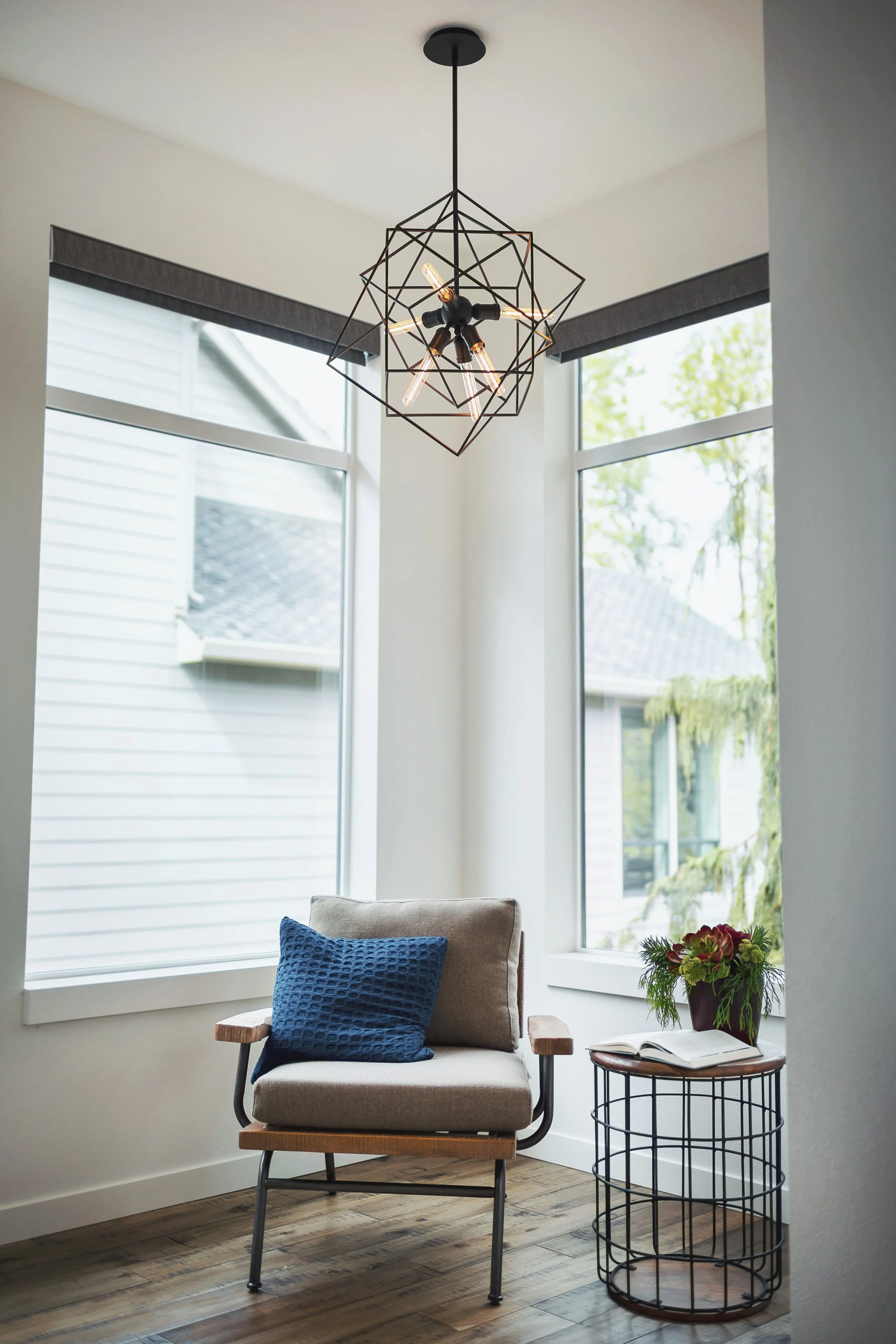 Interior of a cozy living room with large windows, a beige armchair with a blue cushion, a small side table with a potted plant, and a modern geometric chandelier hanging from the ceiling.