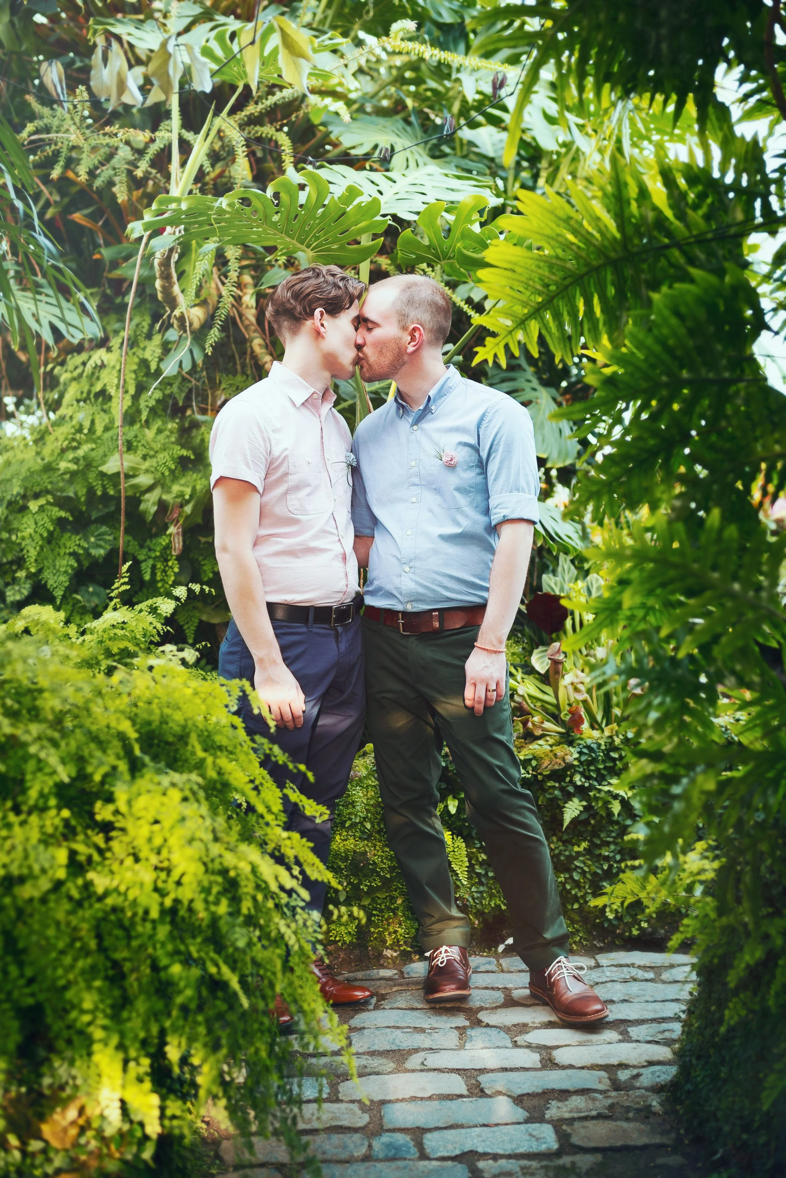 Two men kissing in a lush green garden with dense tropical plants and cobblestone pathway.
