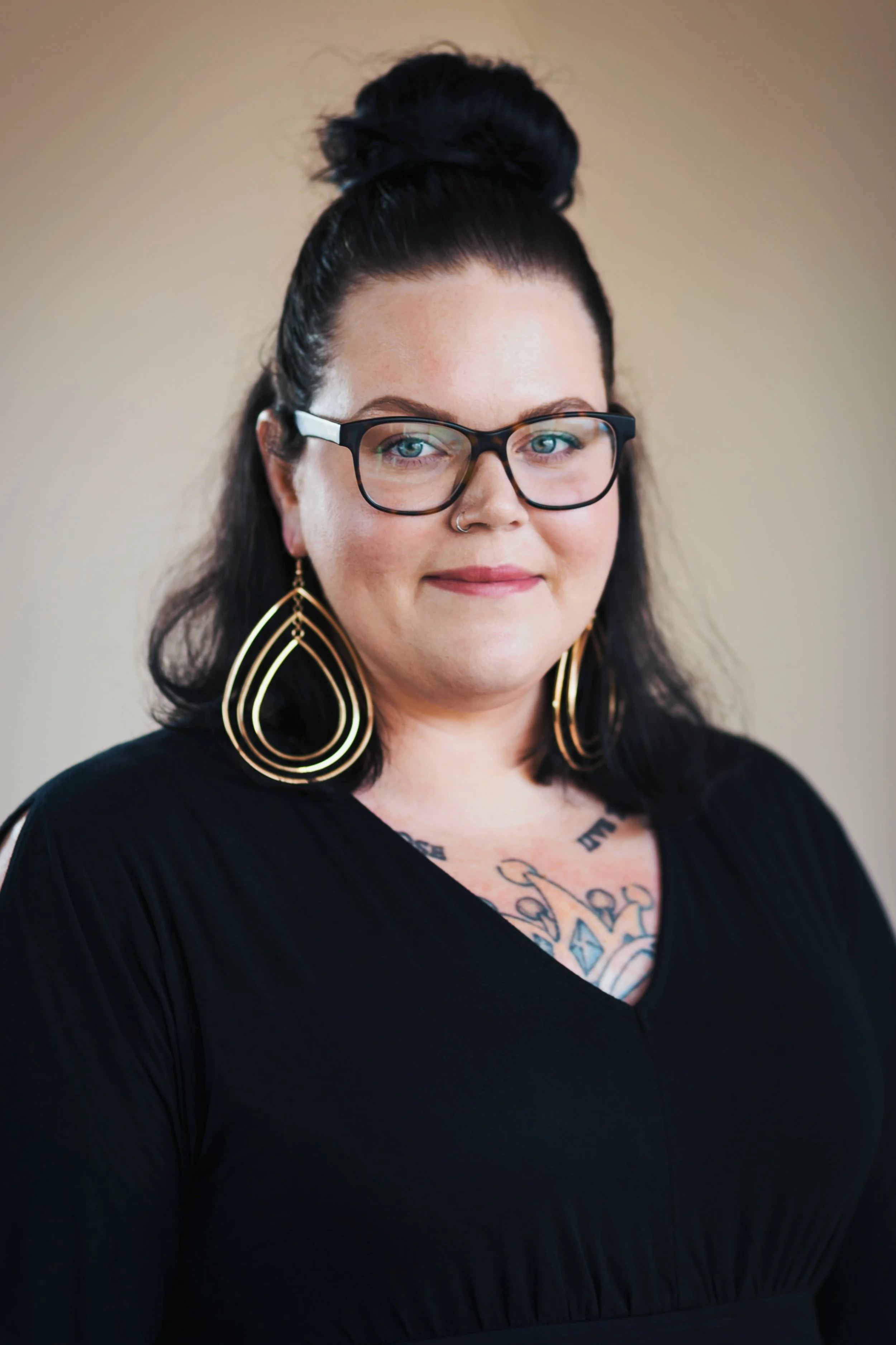 A woman with black hair styled in a top knot, wearing large black glasses, gold earrings, and a black top, standing against a neutral background.