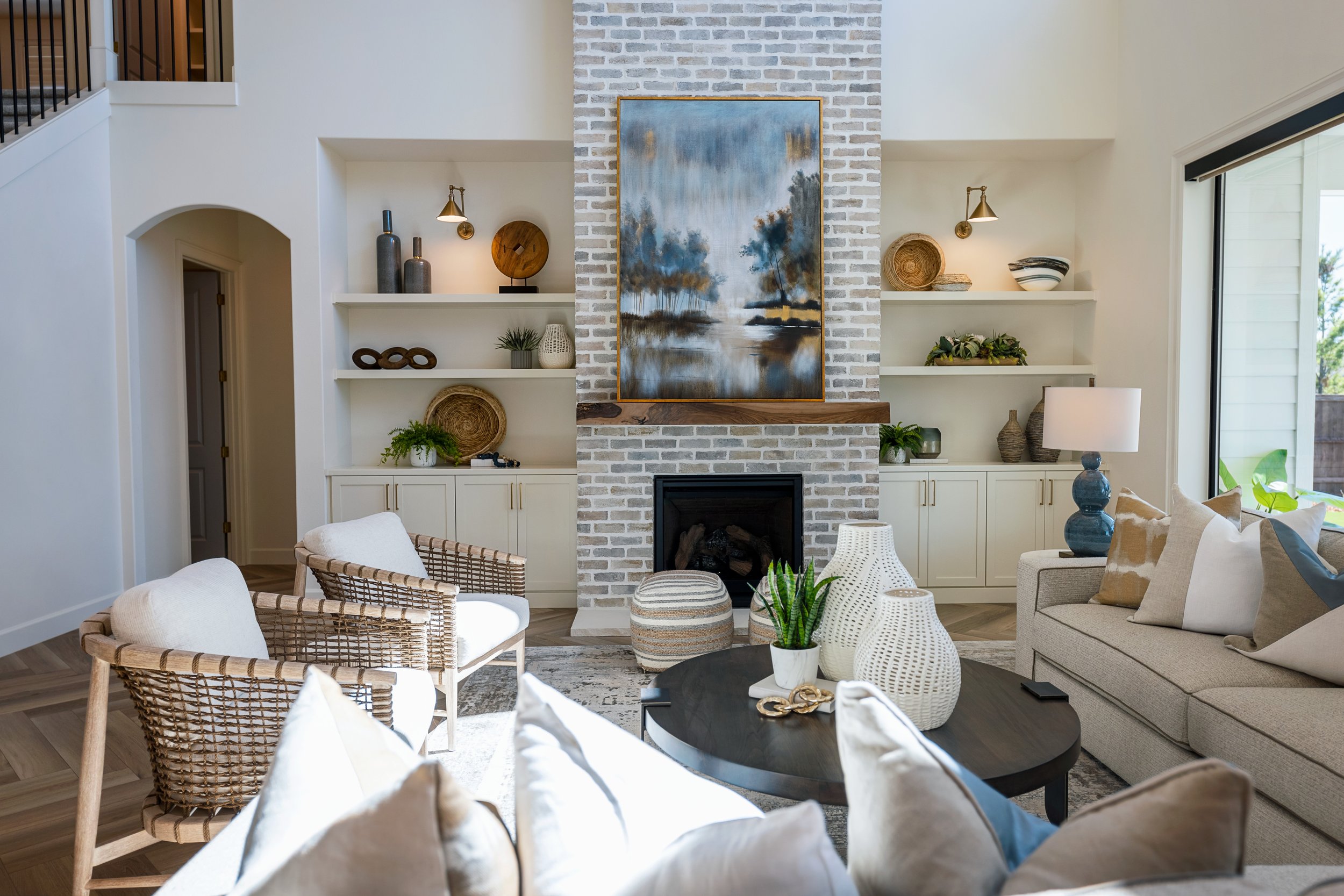 Living room with a white brick fireplace, abstract landscape painting above, built-in shelves with decorative items, and seating area with woven chairs, a sofa with pillows, and a round black table.