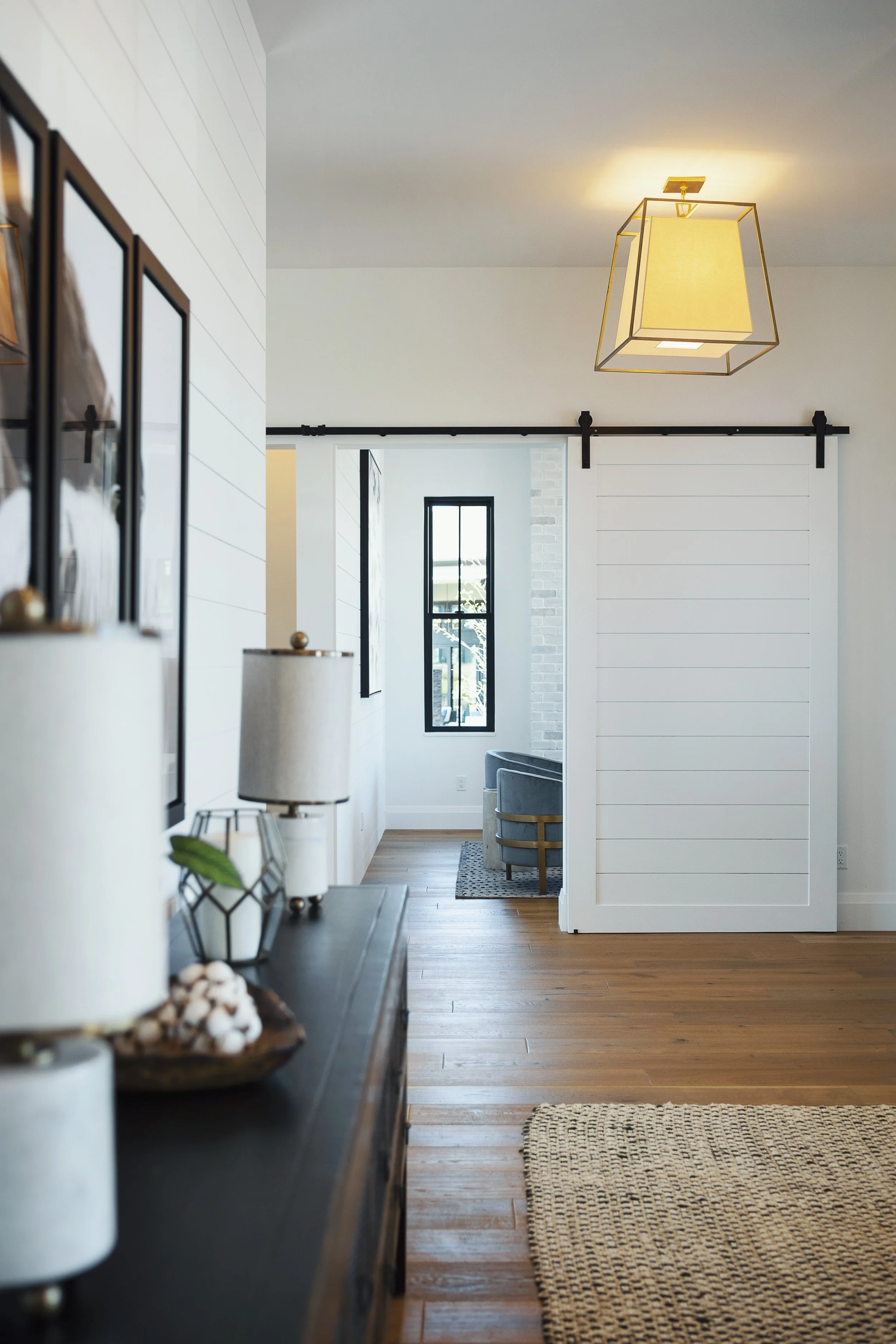 Interior of a home hallway with wood flooring, white walls, black-framed windows, a white barn-style sliding door, and a modern hanging ceiling light.