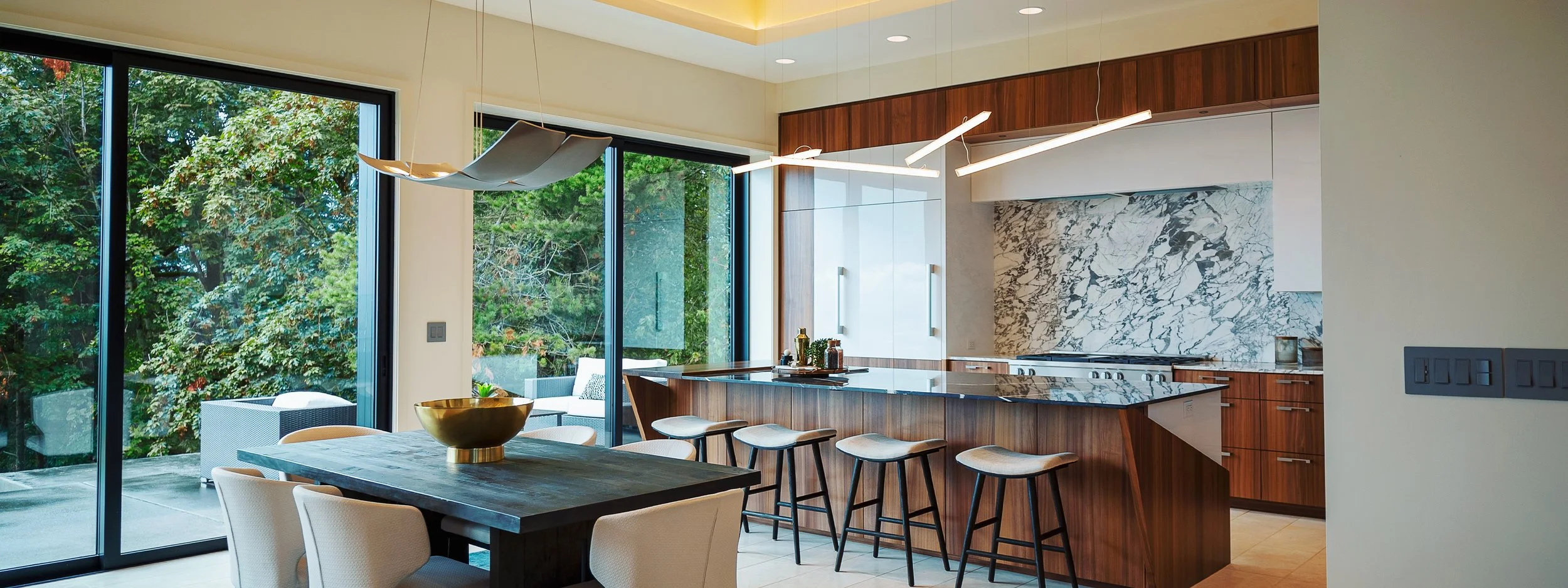Modern kitchen with wooden cabinets, marble backsplash, and a large marble island with bar stools. Sliding glass doors open to an outdoor patio with greenery.