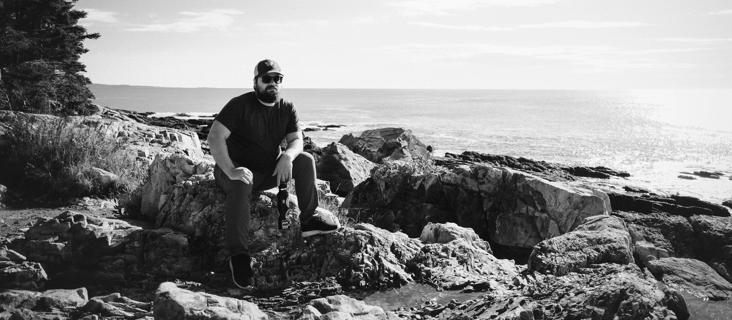 A man with a beard and sunglasses sitting on rocks by the ocean, holding a bottle, wearing a cap, t-shirt, and pants, with trees on the left and the ocean and sky in the background.