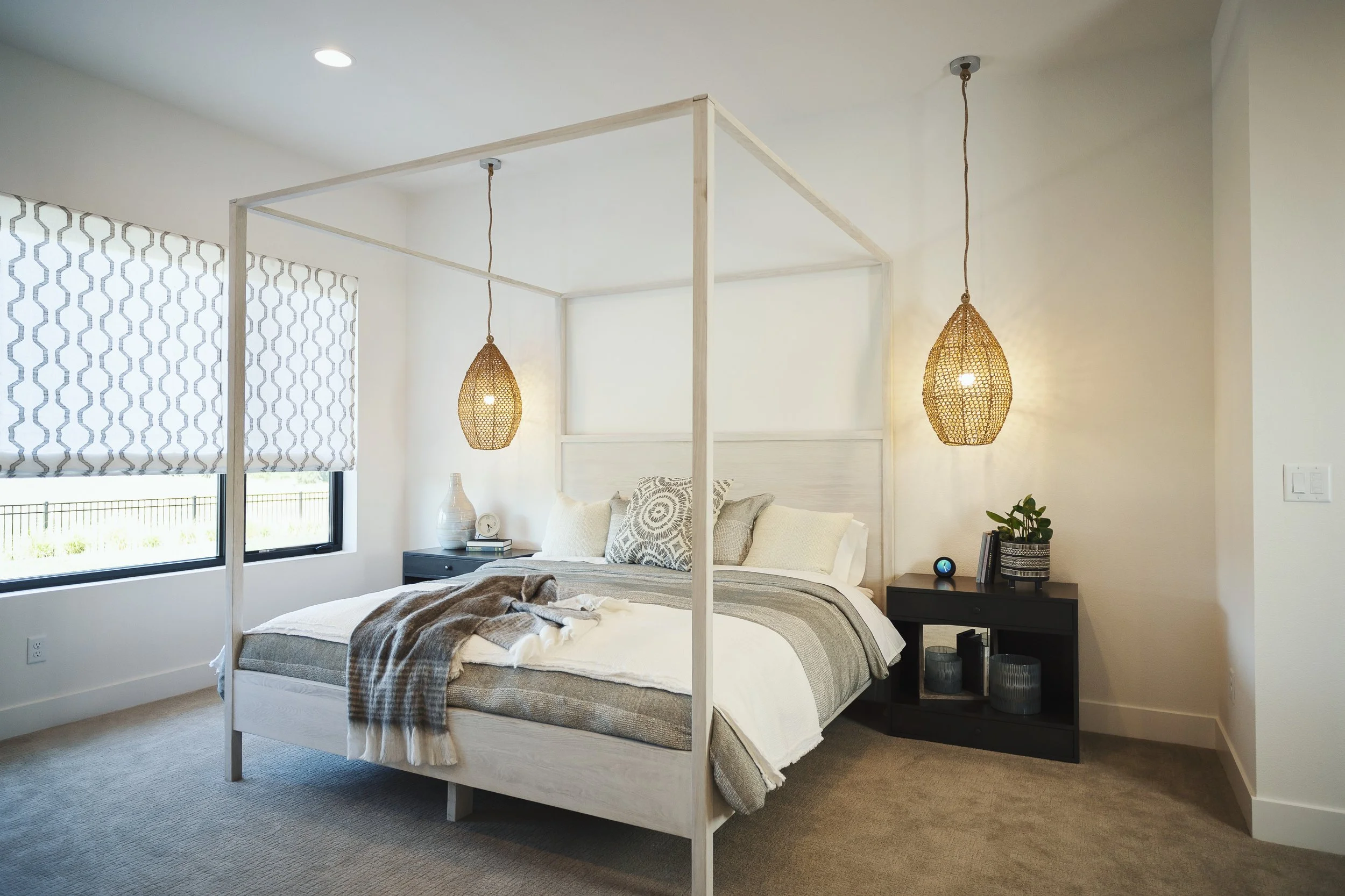 A bedroom with a white bed frame and a bed made with pillows and blankets, two hanging wicker lights, a window with patterned Roman shade, and a small black nightstand with decor items.