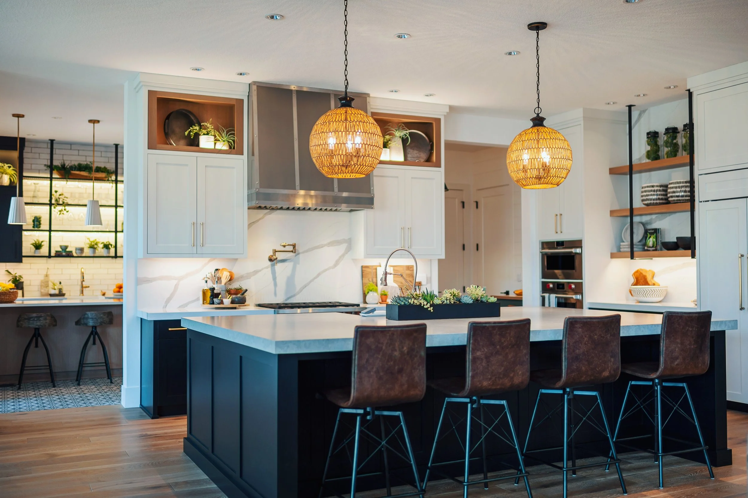 Modern kitchen with white cabinets, a large kitchen island with a dark base, and pendant lights hanging above. Open shelving with glass jars, plants, and decorative items are on the walls, with hardwood floors and a patterned rug.