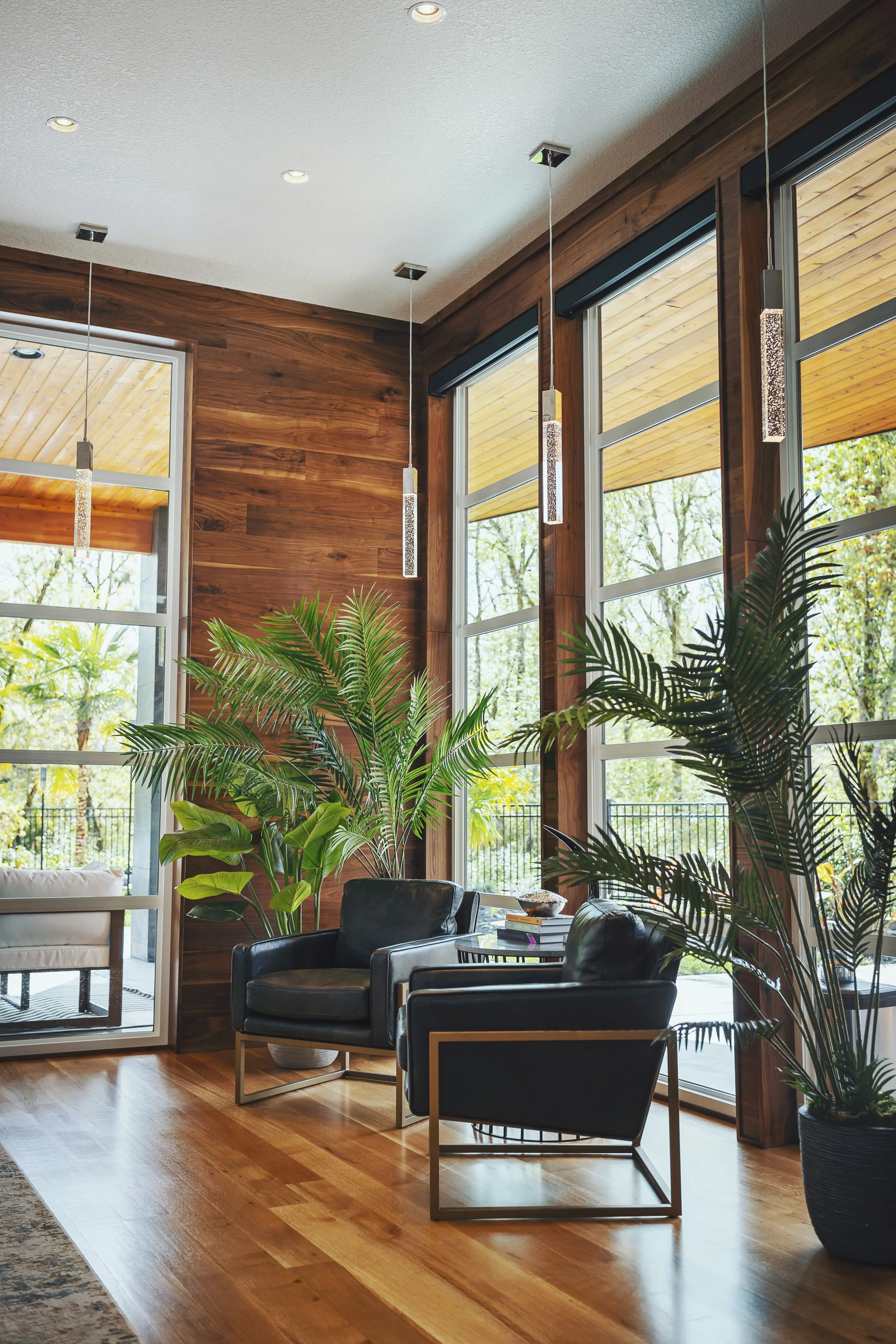 Living room with large windows, wooden wall, black leather chairs, indoor plants, and pendant lighting.