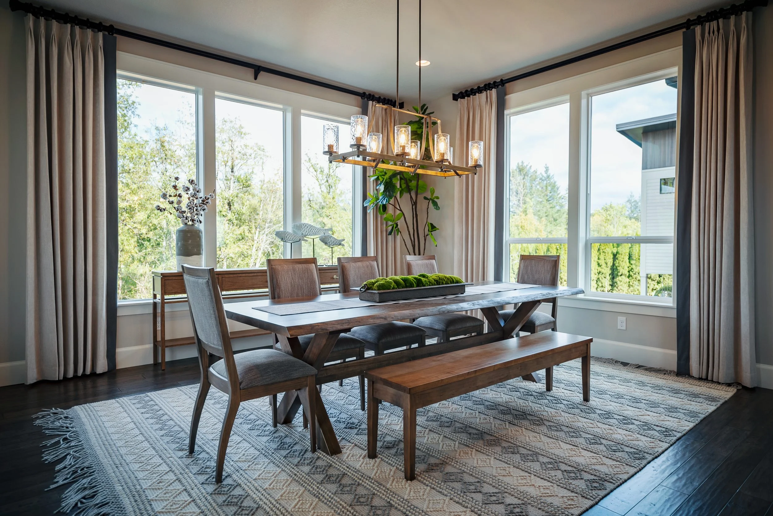 A dining room with a wooden table and six chairs, large windows with beige curtains, a modern chandelier, a rug, a white vase with flowers, and views of greenery outside.