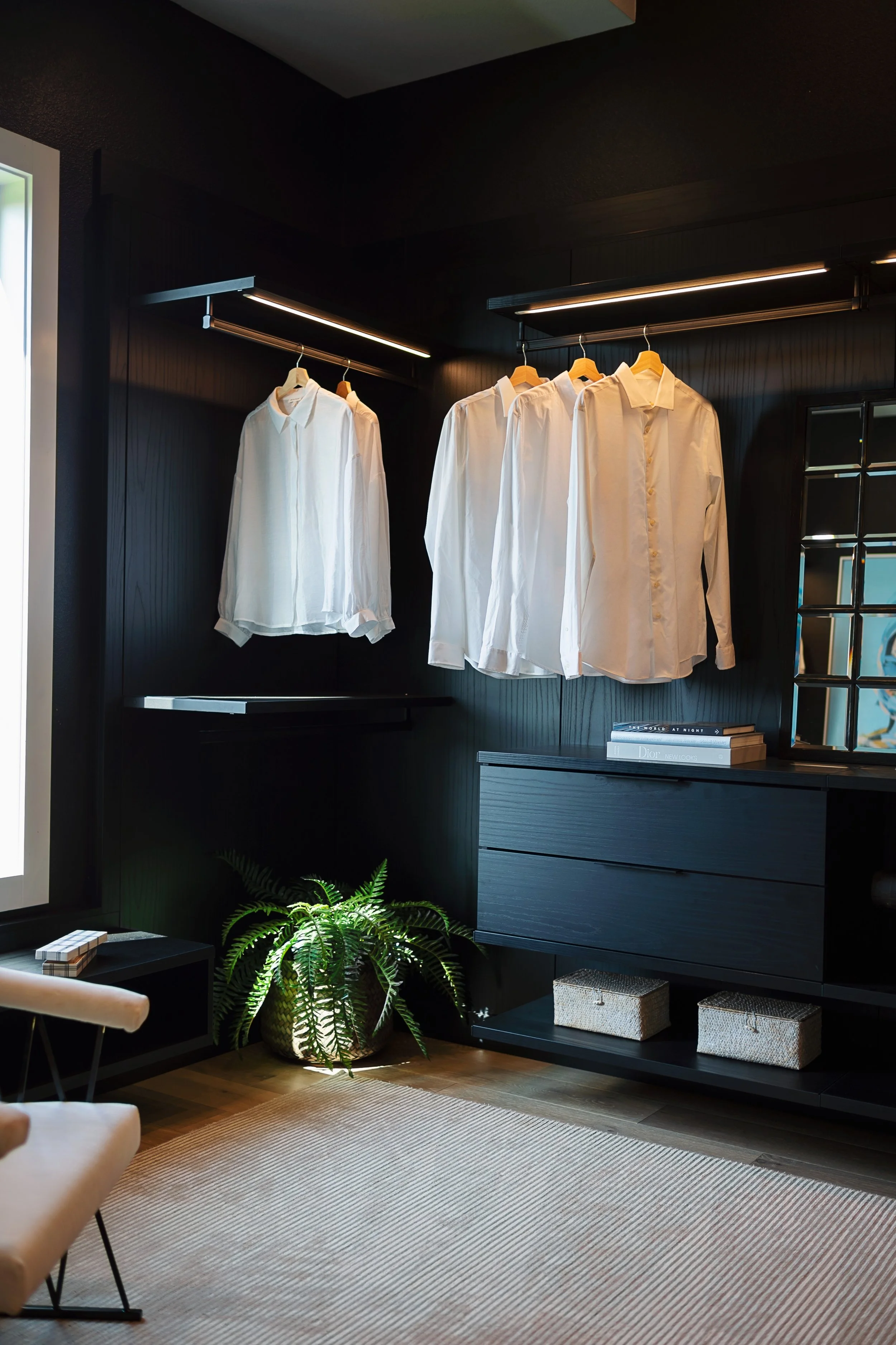Modern black closet with hanging white dress shirts, a potted fern, a beige chair, a black side table, and woven storage baskets on a beige striped rug.