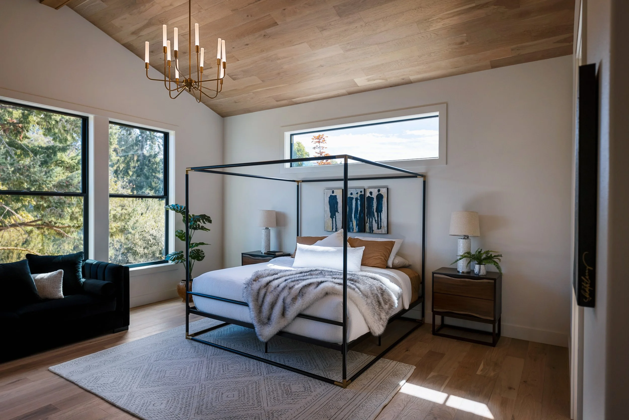 Modern bedroom with a white bed, black metal frame, and two nightstands with lamps. Large windows provide a view of trees and blue sky.