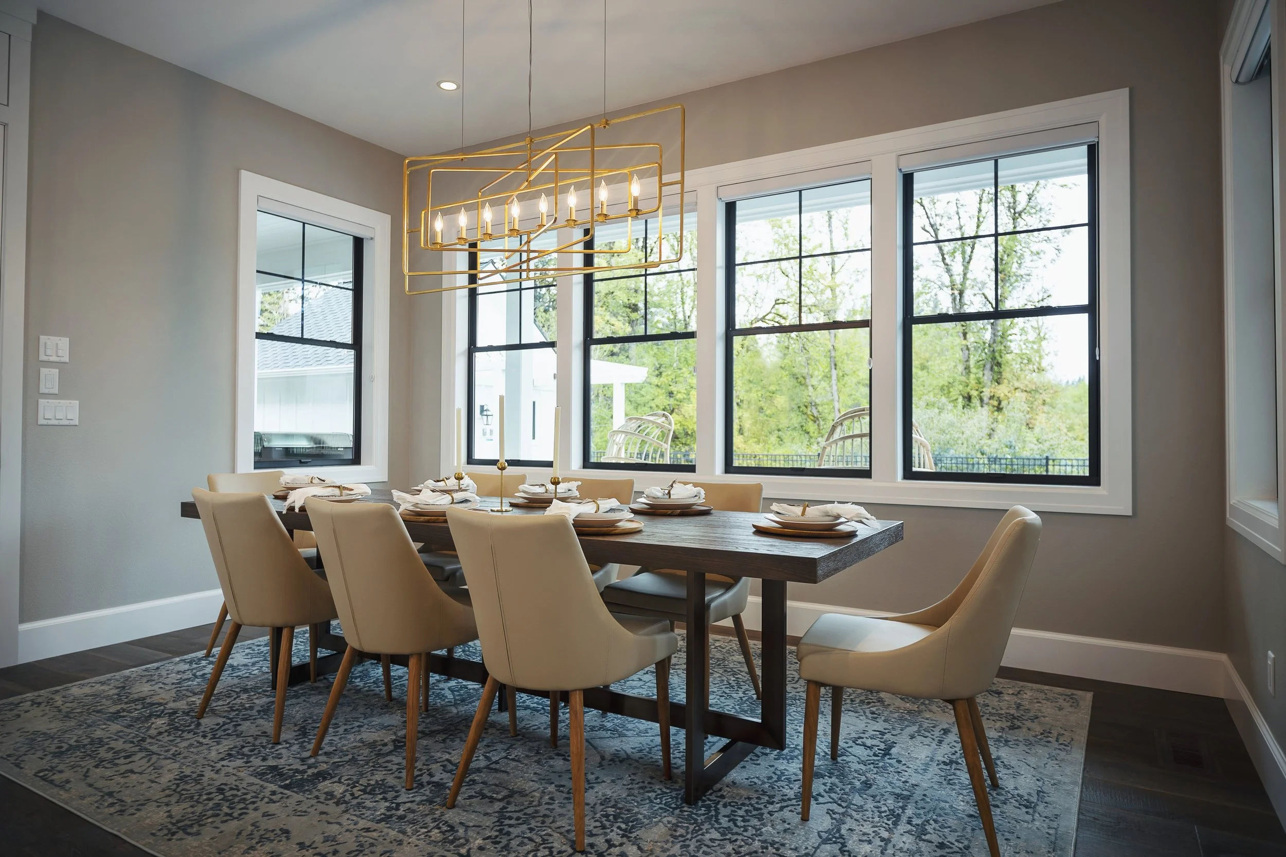 Dining room with a wooden table set with white plates, napkins, and gold-colored candle holders. Beige chairs surround the table. Large windows with black frames let in natural light and show trees outside. A modern gold chandelier hangs above the ta