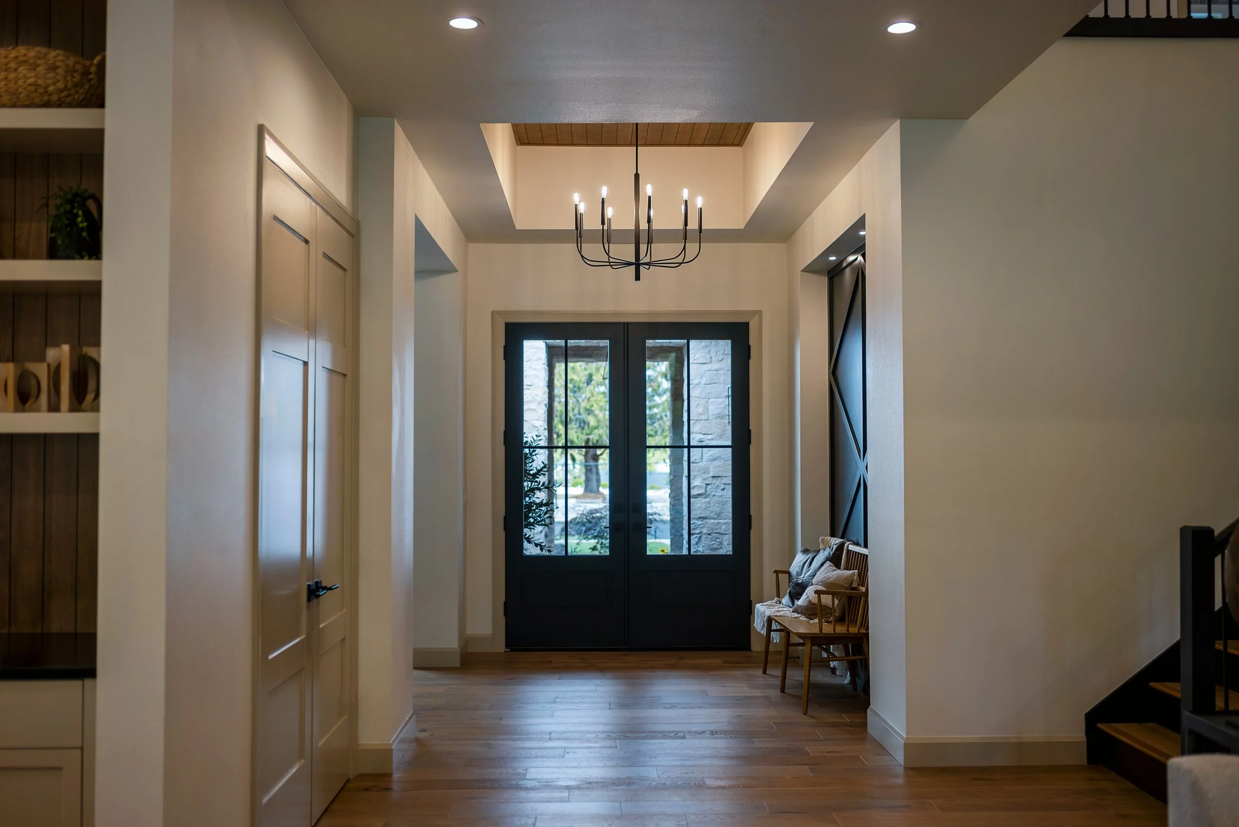 Interior view of a modern home entrance with black double doors and large glass panes, wooden flooring, a chandelier hanging from a ceiling with a tray design, and a small sitting area with chairs on the right.