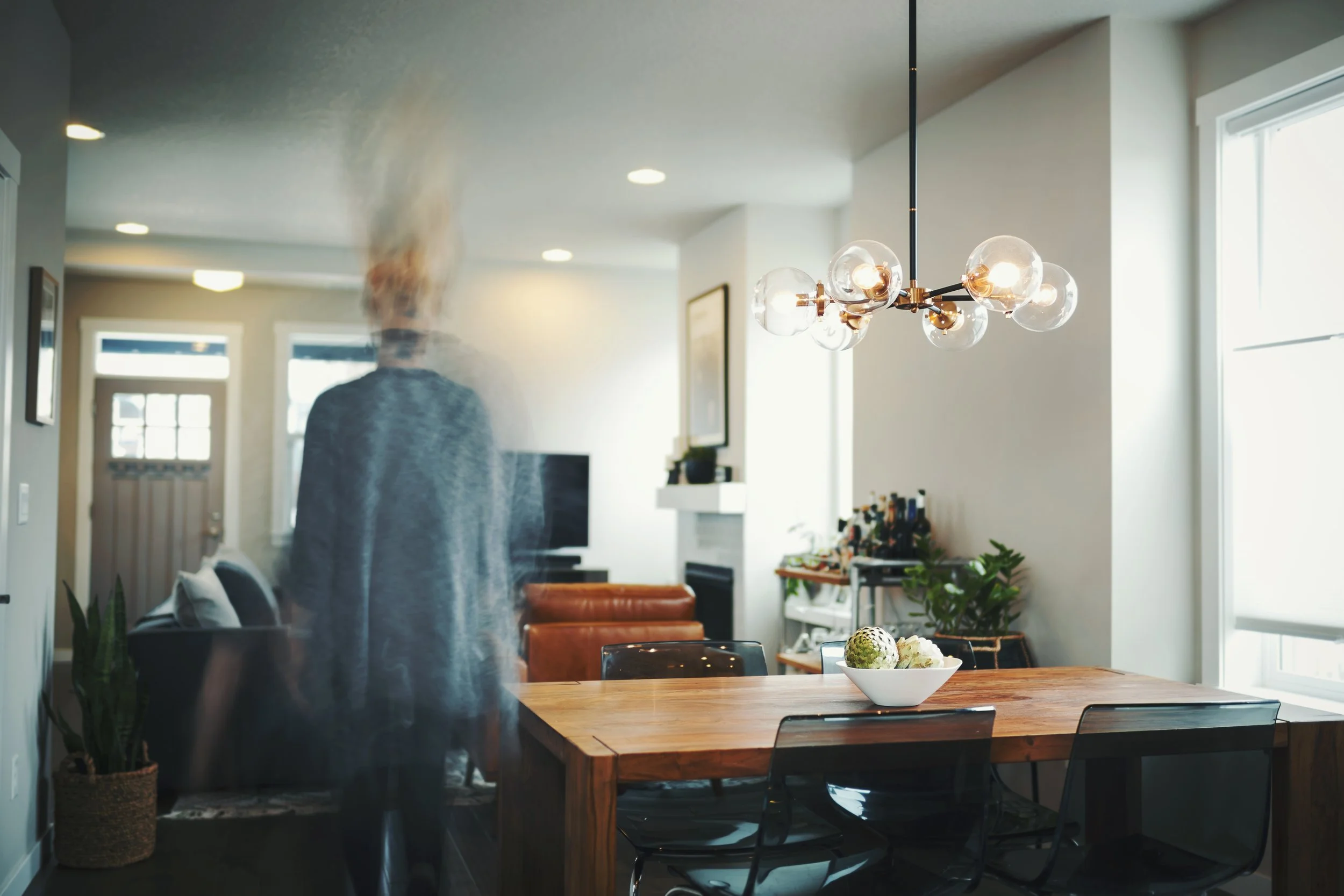 Inside a modern home, a person with gray hair, wearing a gray shirt, is walking past a dining table. The table has a white bowl with decorative objects, and black chairs around it. In the background, there's a bright living space with a fireplace, sh