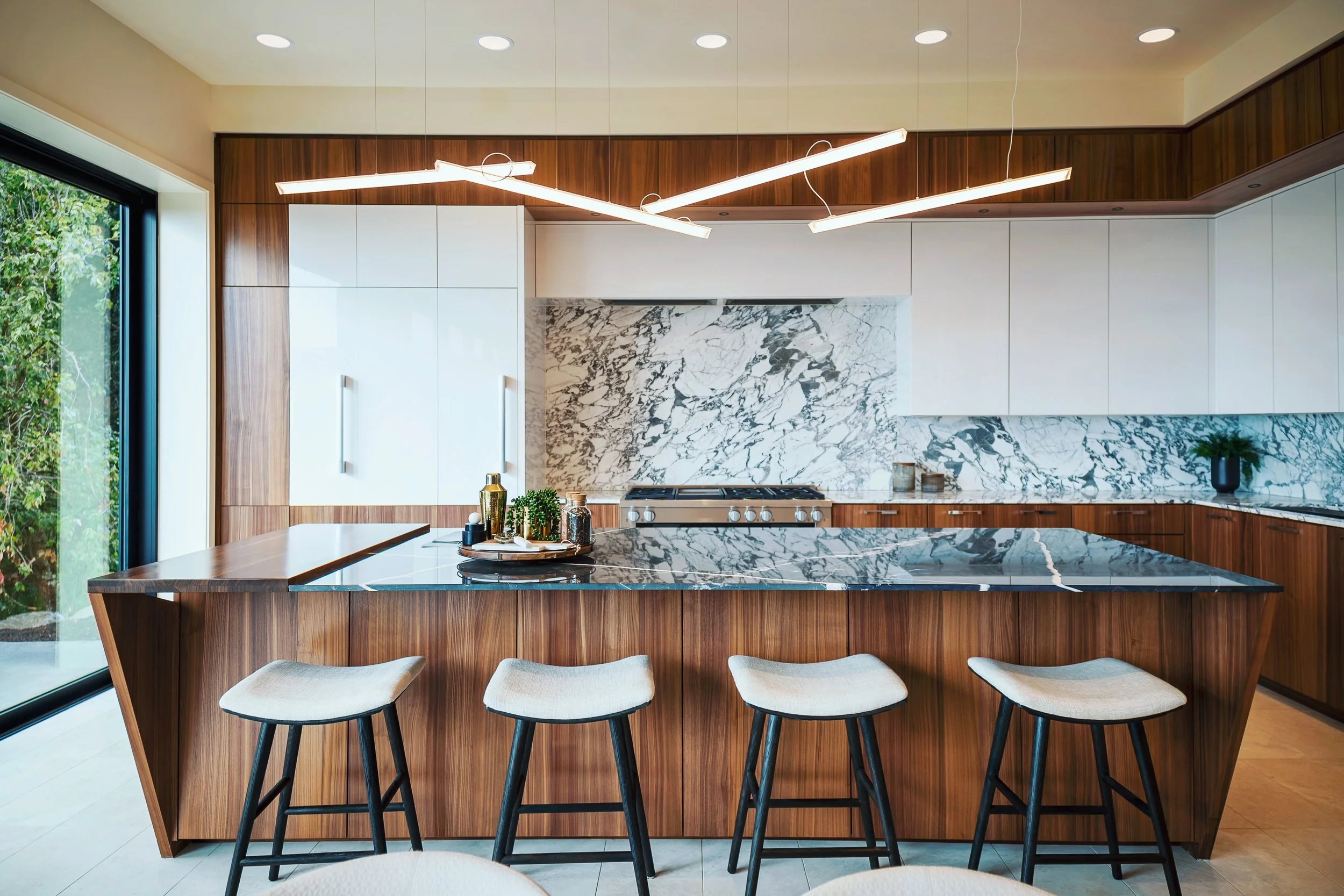 Modern kitchen with wooden cabinets, a white marble backsplash, and a marble island. Four bar stools are placed at the island, and there is a large window showing greenery outside. Contemporary lighting fixtures hang from the ceiling.
