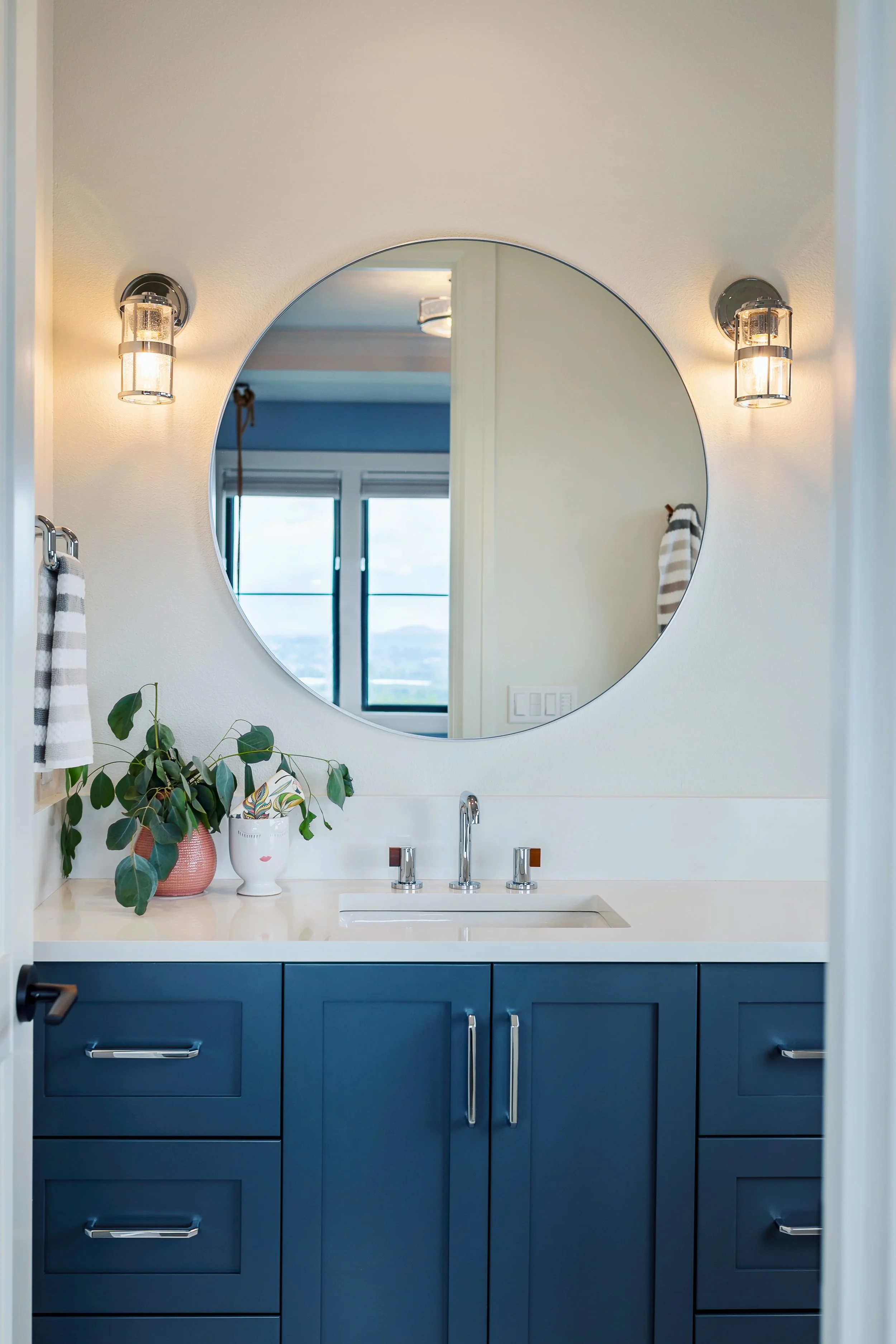 Bathroom vanity with a white countertop, navy blue cabinets, and a round mirror. Two wall-mounted light fixtures and a potted plant and decorative vase on the countertop.