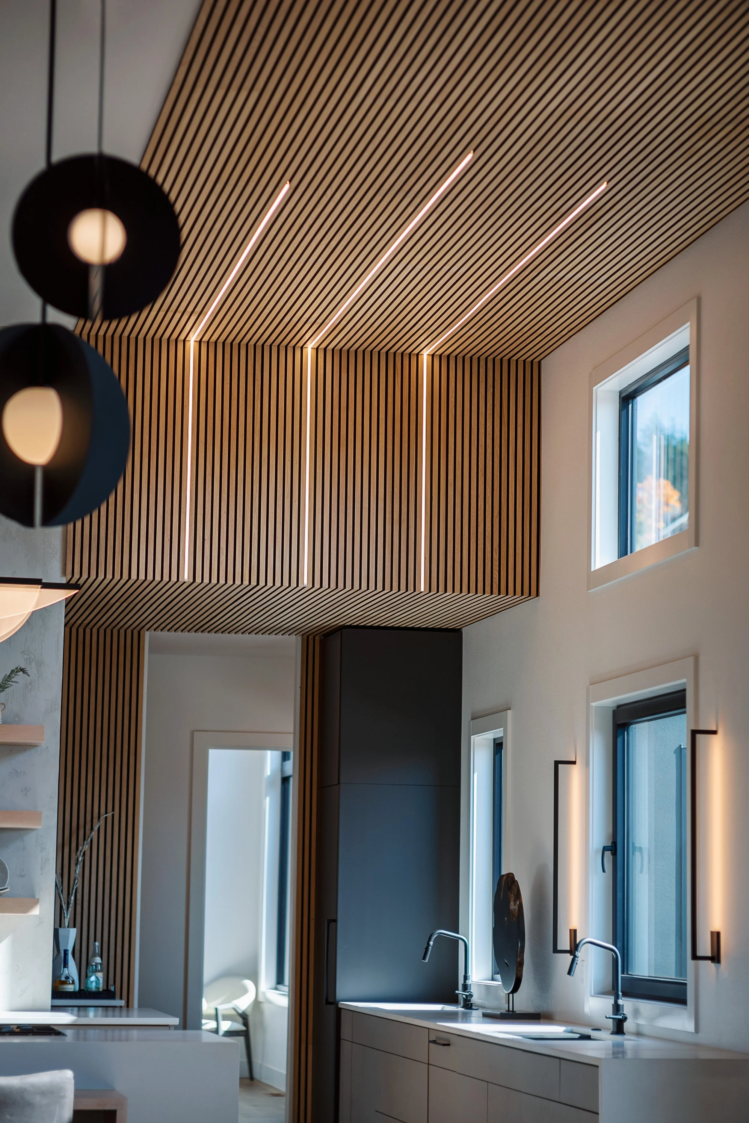 Modern kitchen with wooden slat wall and ceiling, large window with view outside, kitchen sink with faucets, and black pendant lights.