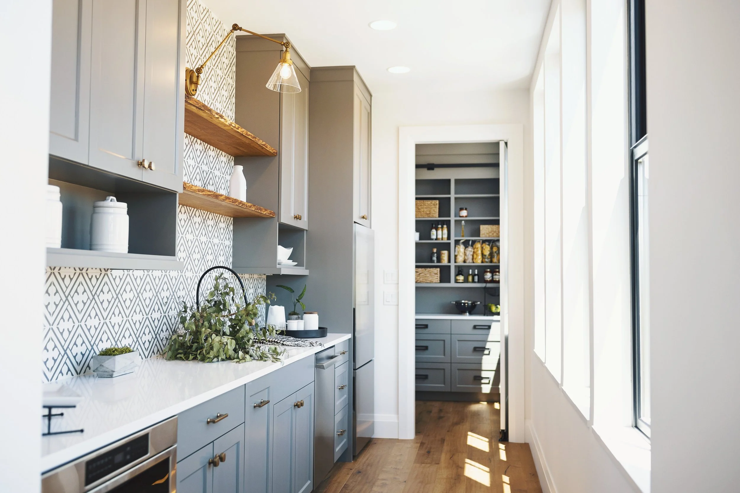 A modern kitchen with light blue cabinets, open wooden shelves, and a decorative backsplash. There are containers on the counter with greenery, and a doorway leads to a pantry with dark shelving and jars.