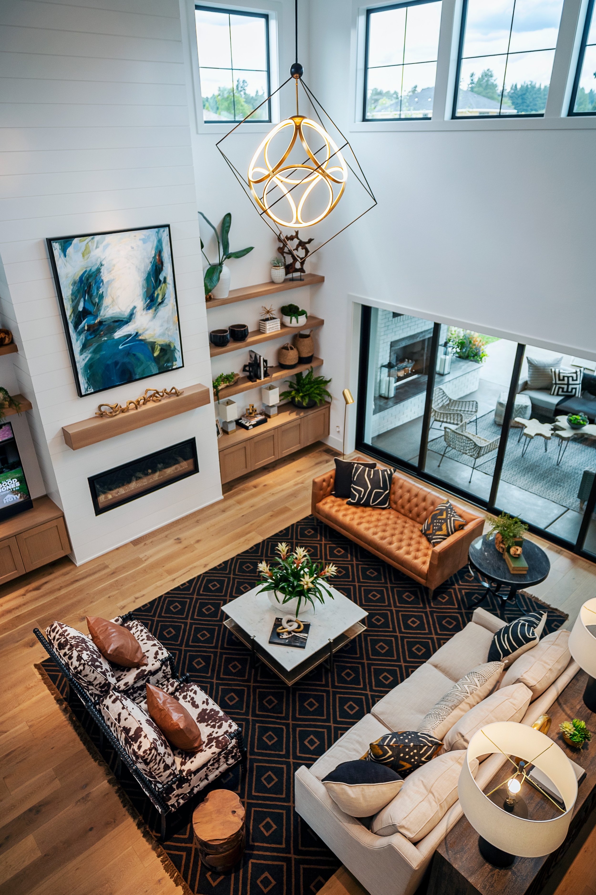 Living room with high ceiling, large windows, modern chandelier, abstract artwork, multiple sofas, chairs, coffee table with plant, and sliding glass doors leading to outdoor patio.