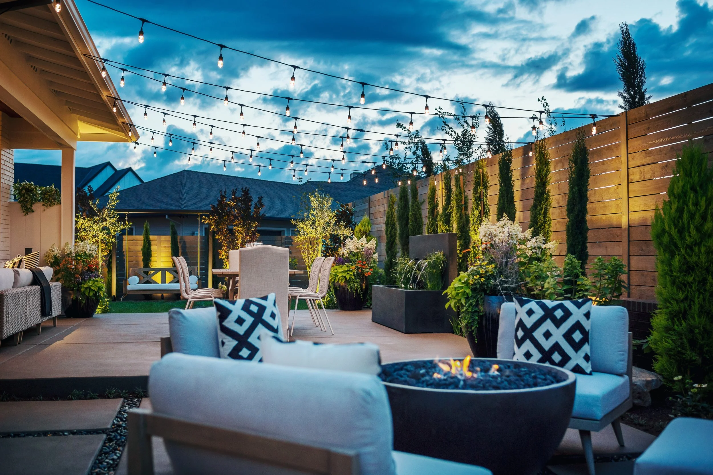 An outdoor patio area at dusk with string lights hanging above, surrounded by a wooden privacy fence and lush greenery including young trees and potted plants. There are patio chairs, a cushioned seating area, and decorative pillows, creating a cozy 