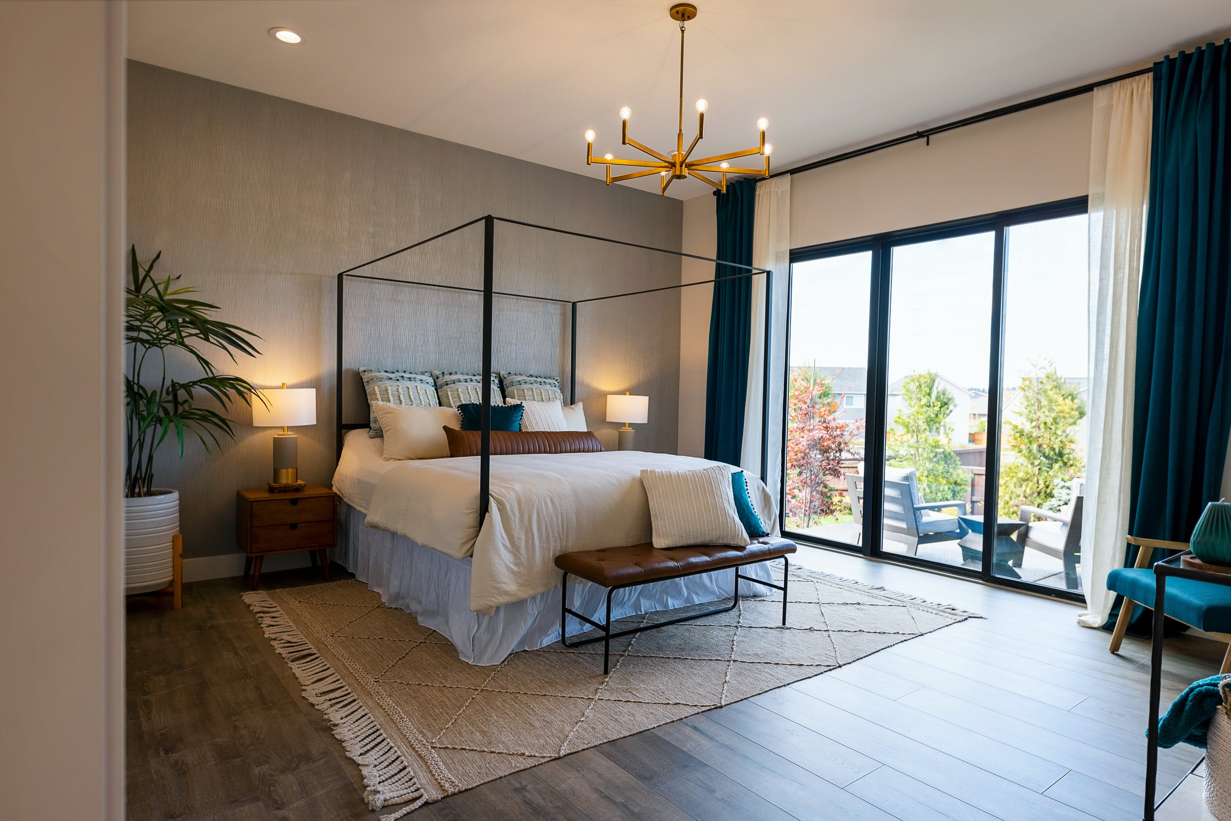 A modern bedroom with a large window overlooking trees, decorated with a bed, lamps, a plant, and a chandelier.
