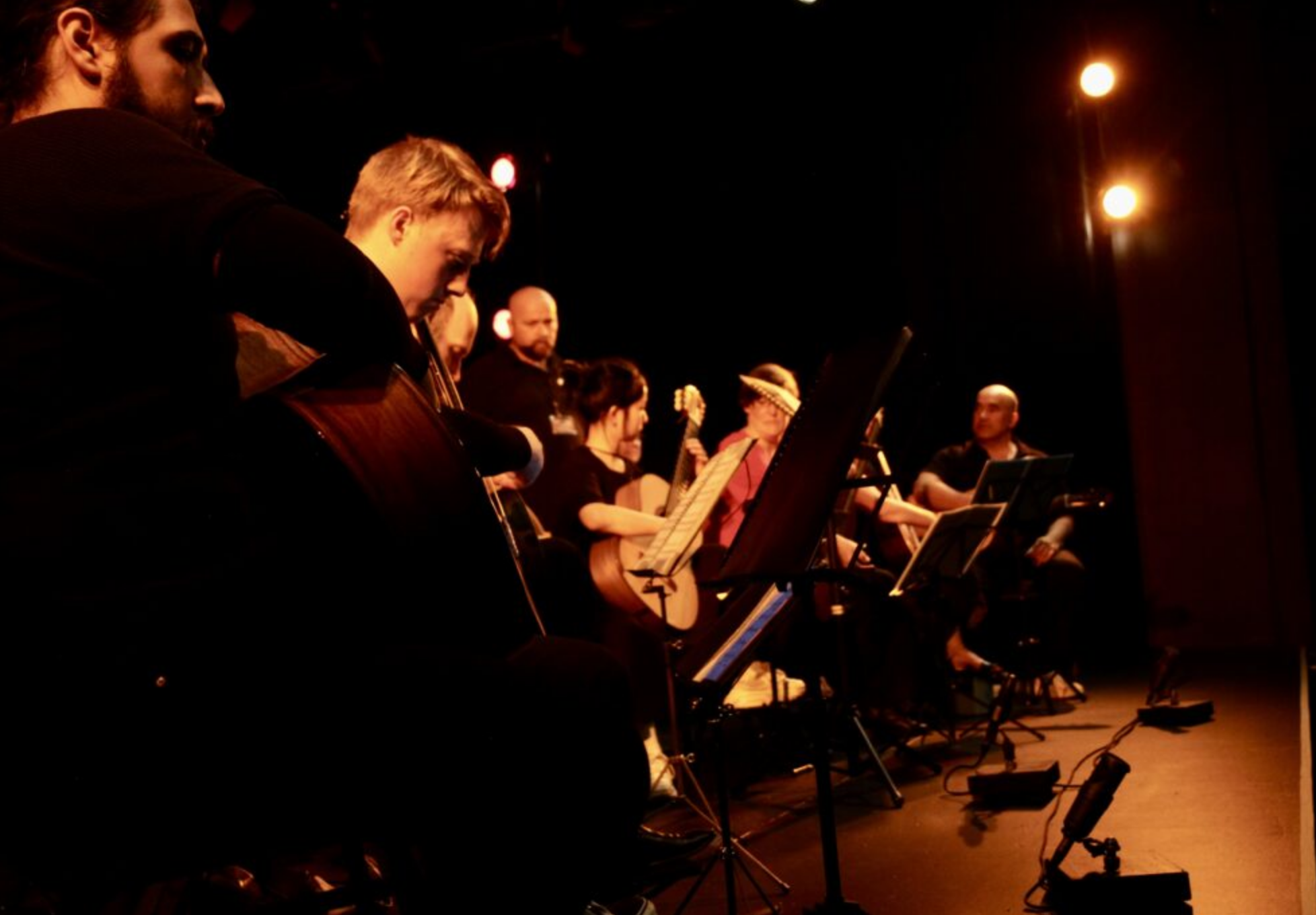 A group of musicians performing on stage, playing guitars and other string instruments, with music stands and stage lights illuminating them.