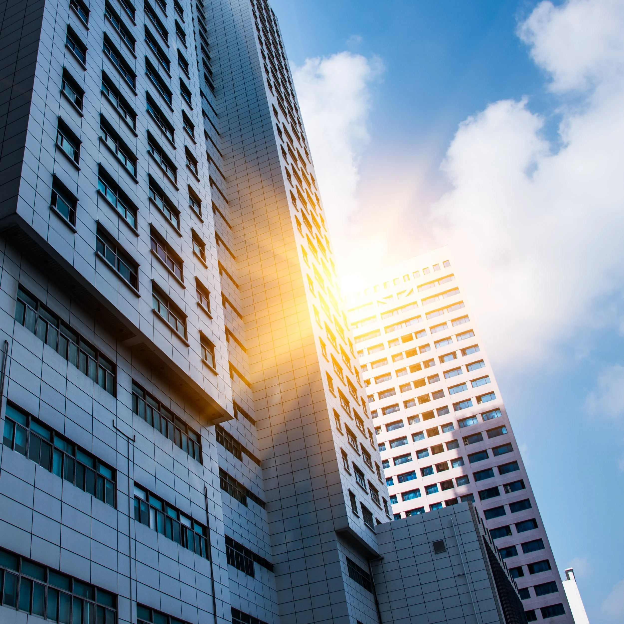 Tall modern skyscrapers with grid windows, sunlight reflecting off the buildings, against a blue sky with clouds.