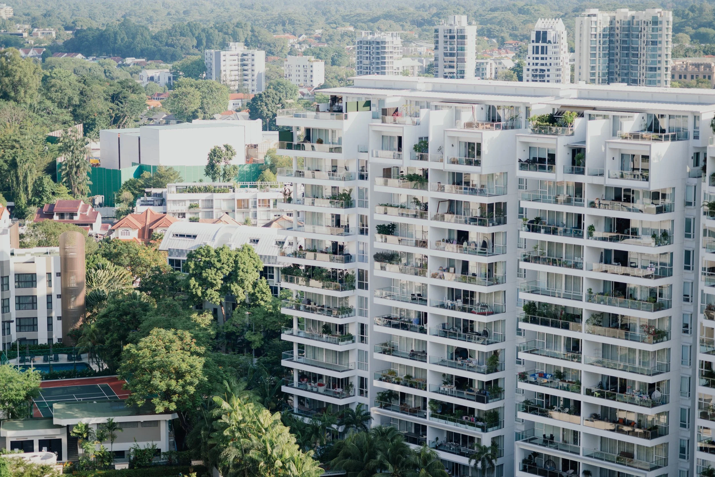 High-rise residential building with multiple balconies, surrounded by trees and other buildings in a cityscape.