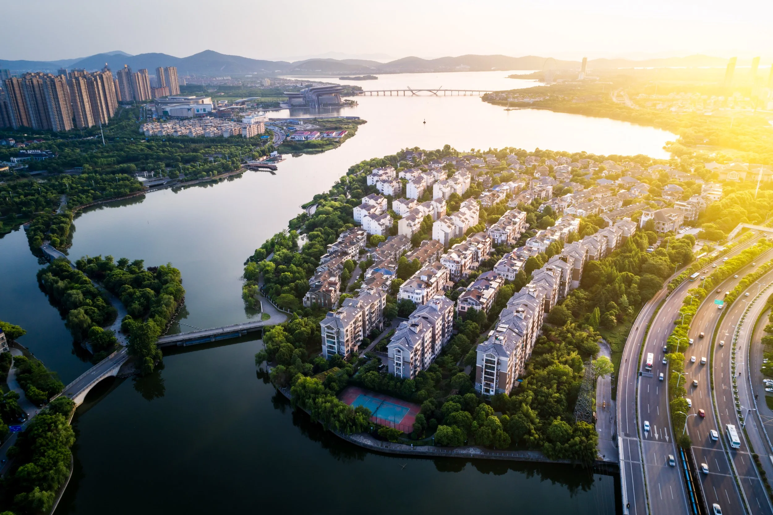 An aerial view of a cityscape during sunset, showing a river with surrounding green parks and residential buildings, with a highway on the right side of the image.