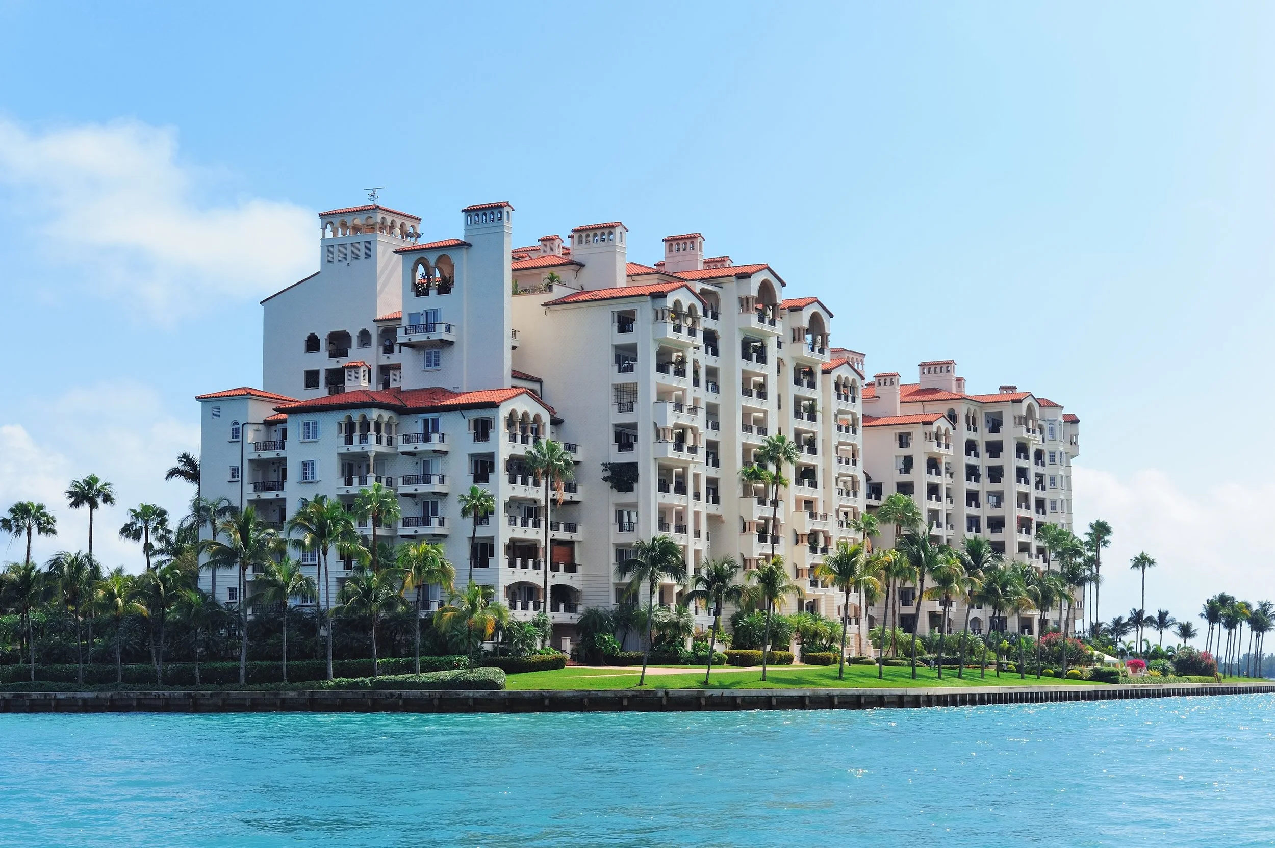 Large white Mediterranean-style hotel with multiple balconies, red-tiled roofs, and palm trees in front, along a waterway under a partly cloudy sky.
