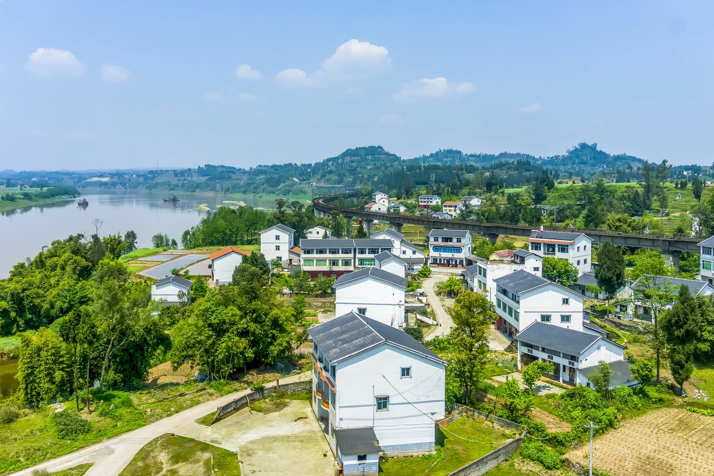Scenic view of a rural village with white houses, green trees, a river, and distant hills under a partly cloudy sky.