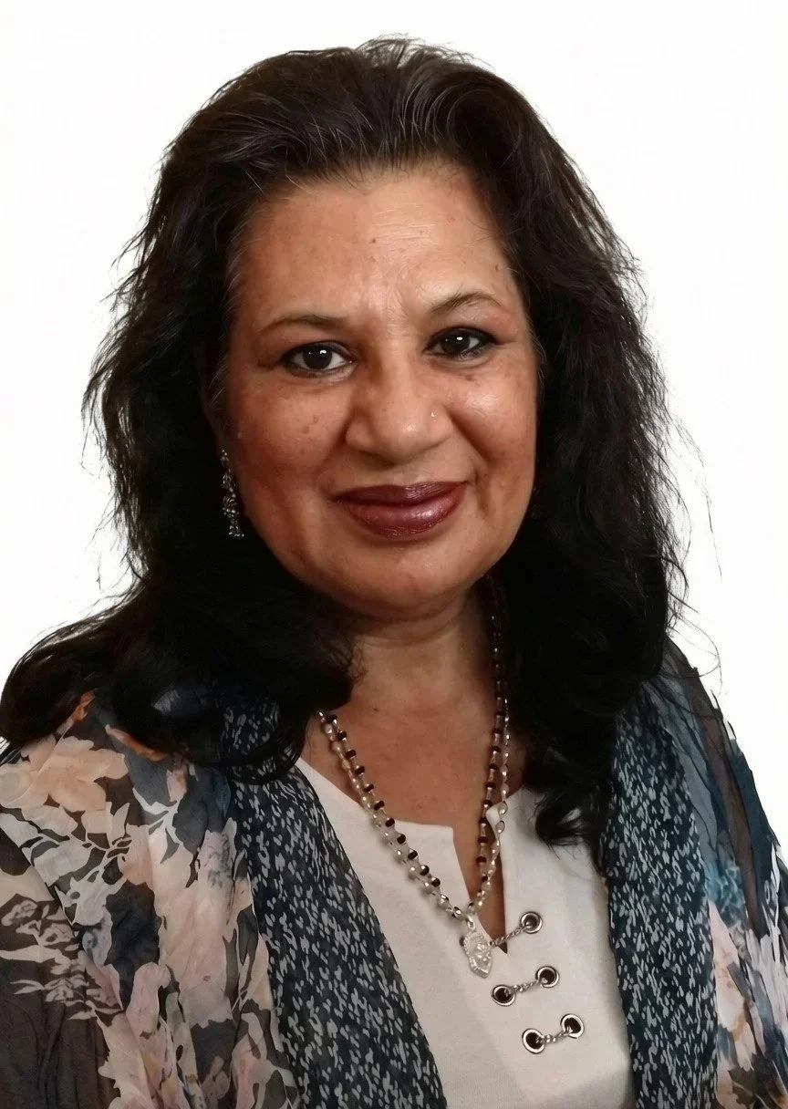 Portrait of a smiling woman with dark hair, wearing a patterned blouse and jewelry, against a plain white background.